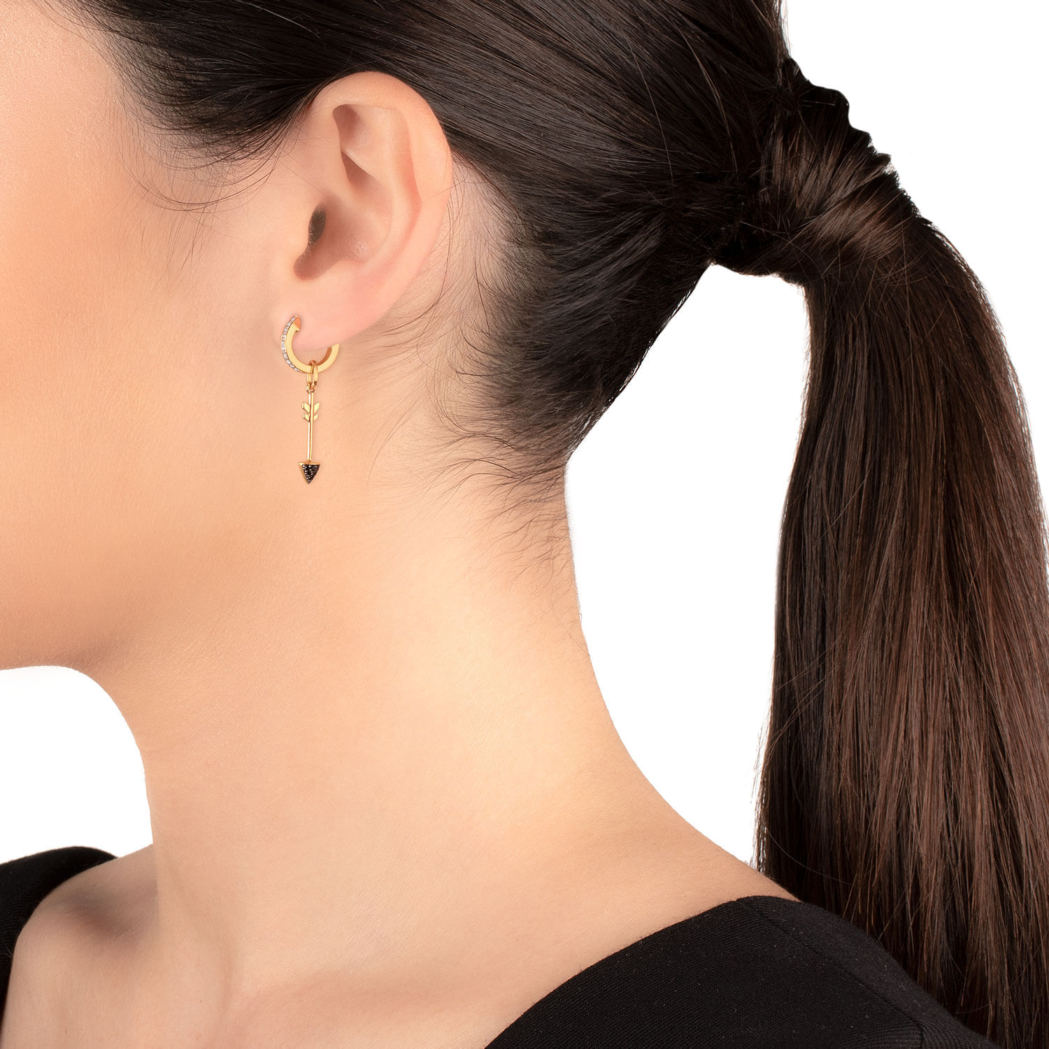 Close da orelha de uma mulher usando um brinco de argola de ouro com um pingente de triângulo preto e um pingente de flecha, com seu cabelo escuro preso em um rabo de cavalo.