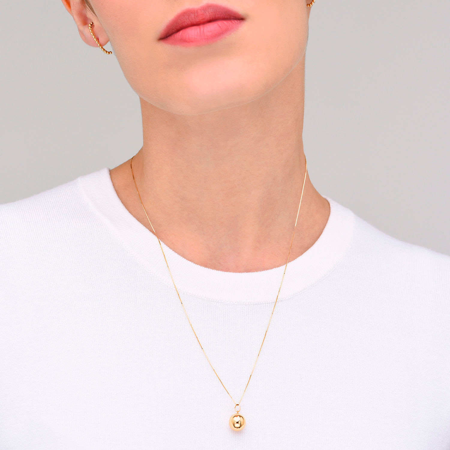 Colar dourado com pingente de esfera, usado por uma pessoa com camiseta branca.