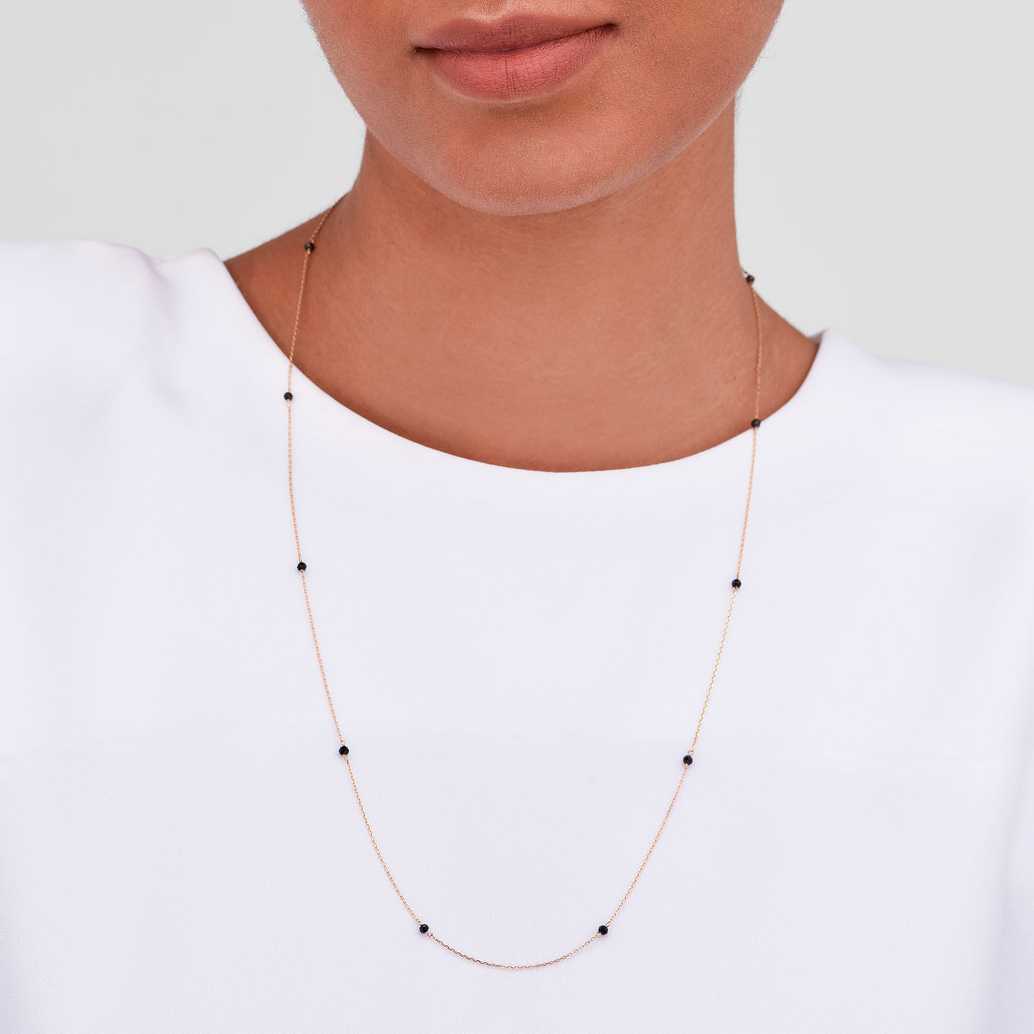 Colar delicado com elos finos e pequenas pedras pretas, usado por uma pessoa com blusa branca.
