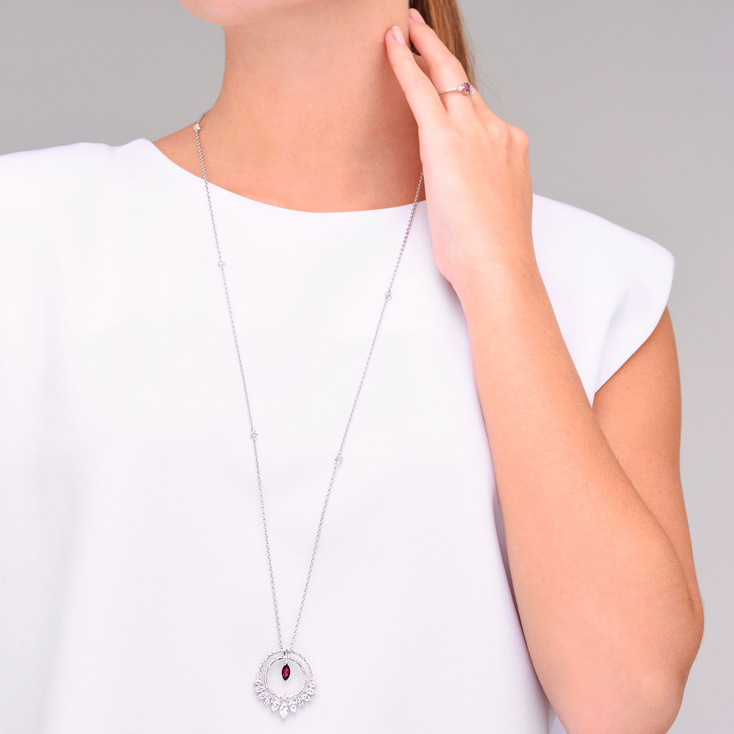 Colar delicado de prata com pingente circular, adornado com pedras transparentes e uma pedra central vermelha, usado com uma mão tocando o pescoço.