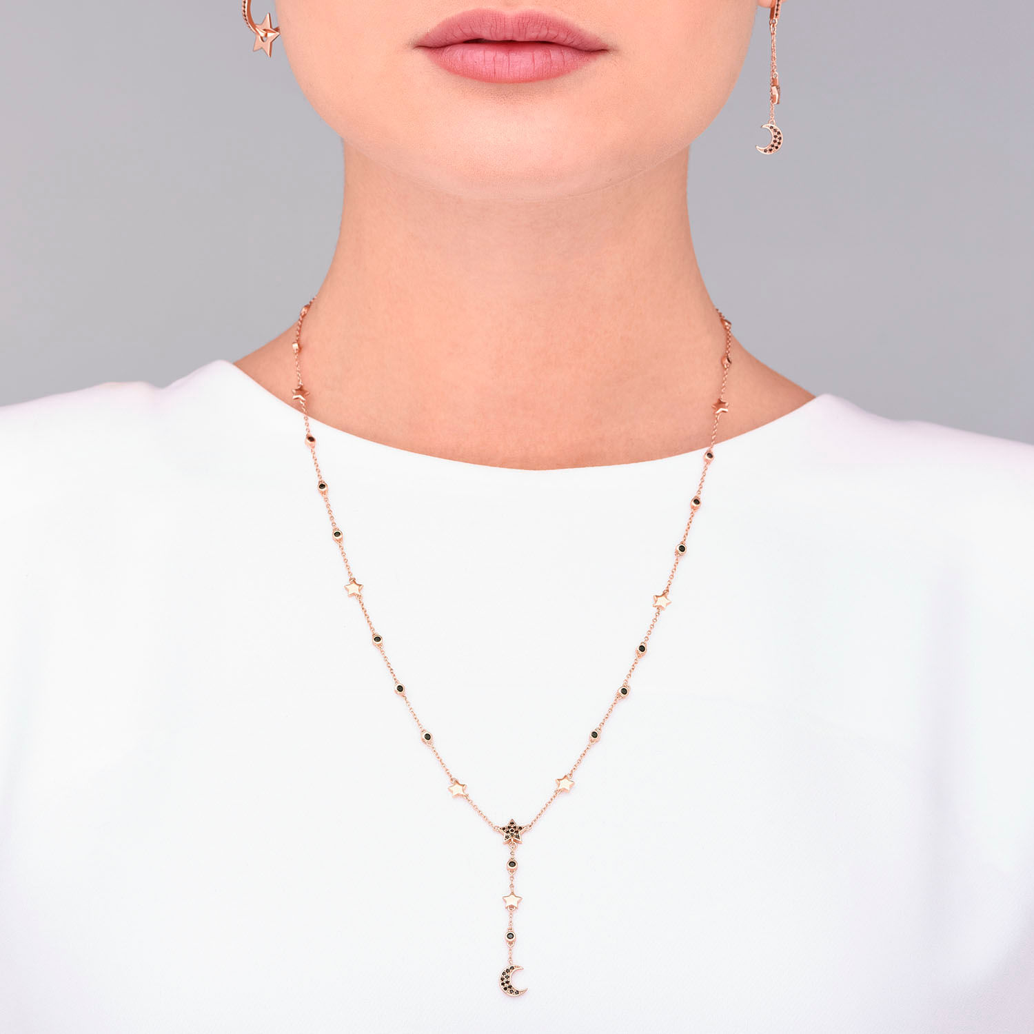 Colar e brincos delicados em tom rosé com detalhes de lua e estrelas, usado por uma pessoa com blusa branca.