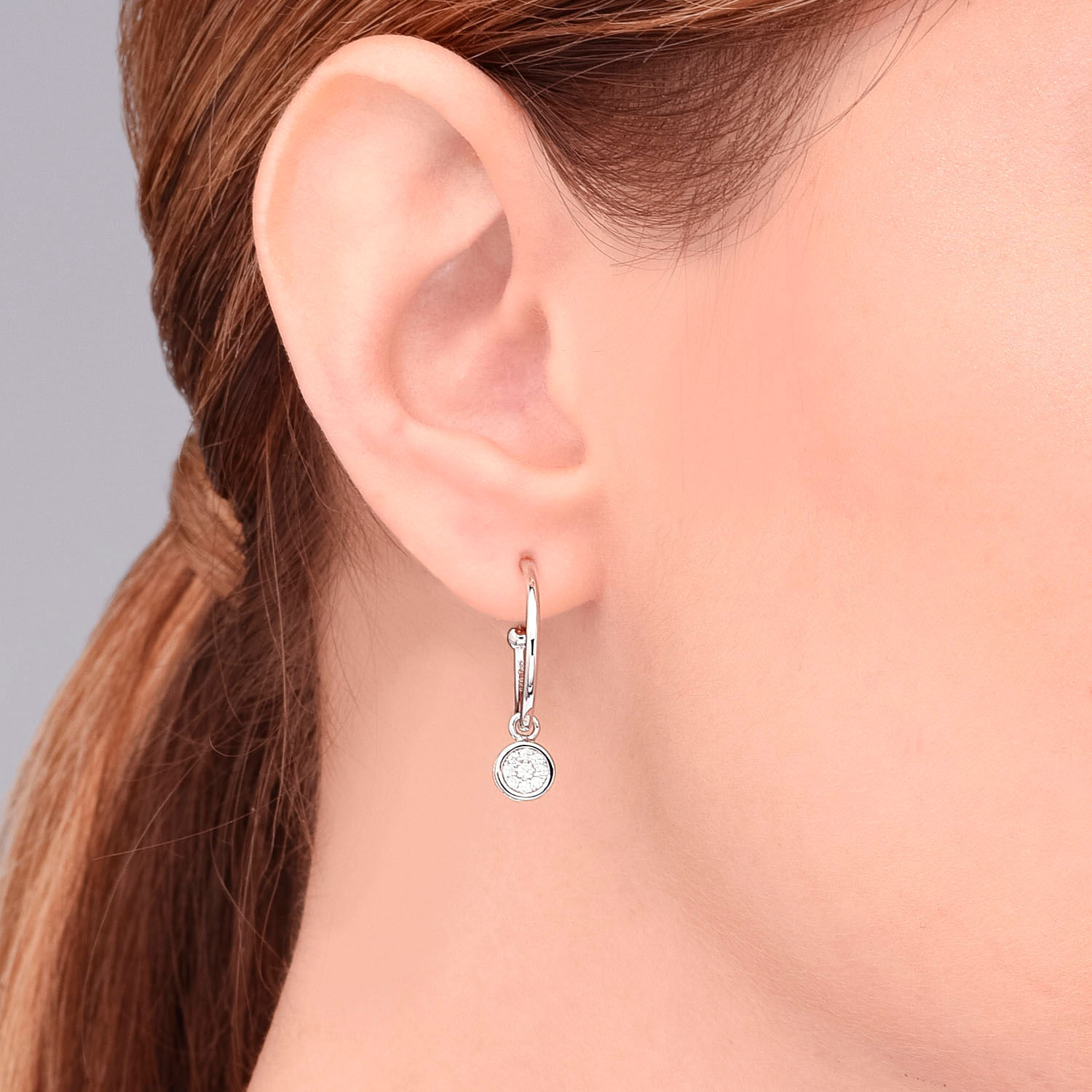 Close da orelha de uma mulher usando um brinco de prata com uma pequena pedra preciosa redonda e transparente.