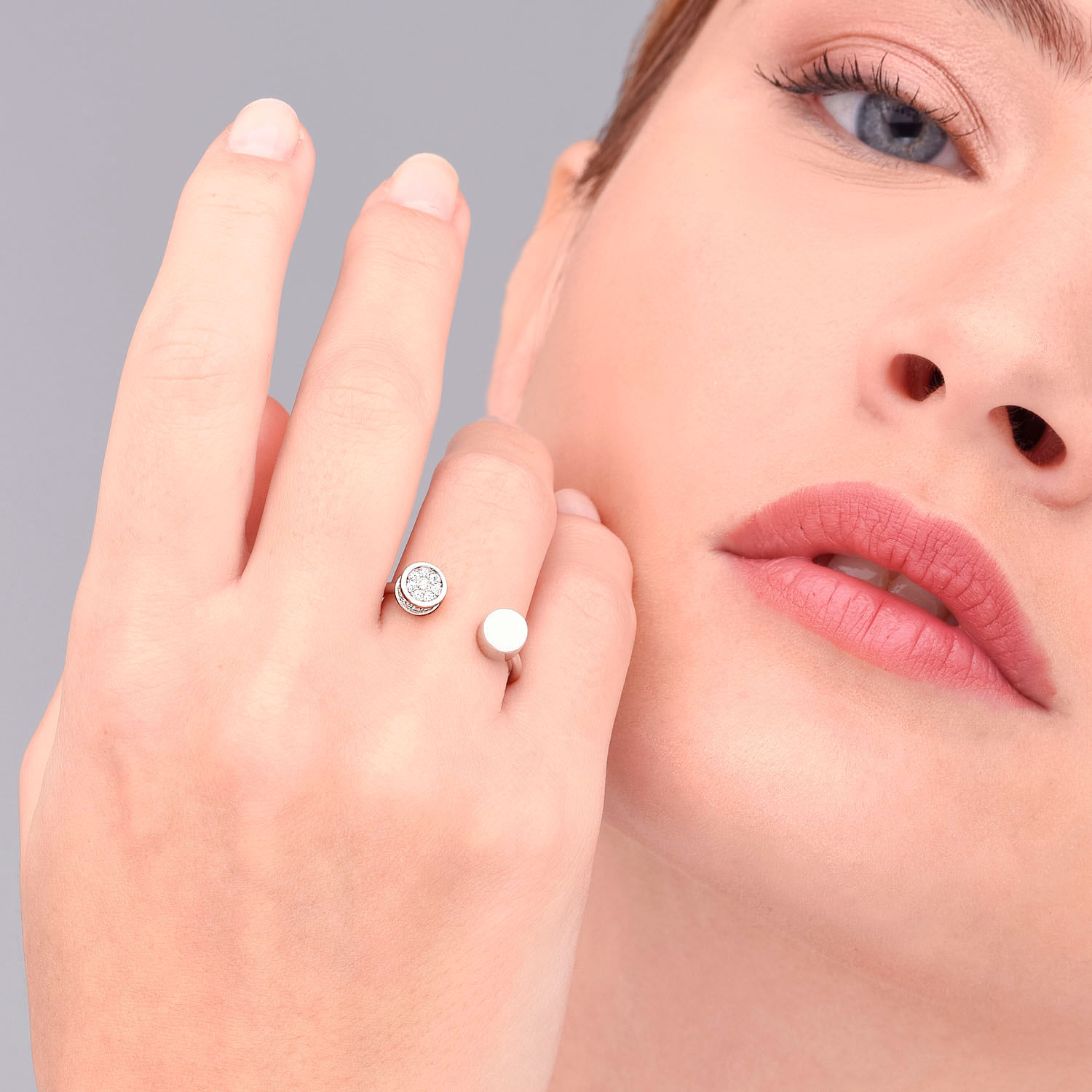 Close-up de uma mulher com pele clara, usando dois anéis finos na mão esquerda, uma com diamantes e a outra branca, perto do rosto.