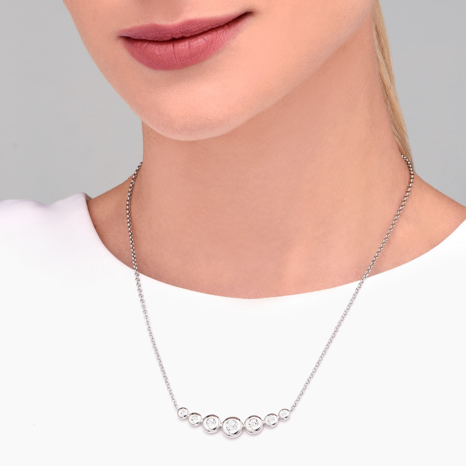 Colar prateado com sete bolinhas de diferentes tamanhos, decoradas com pedras brilhantes, usado por uma mulher com lábios rosados e camiseta branca.