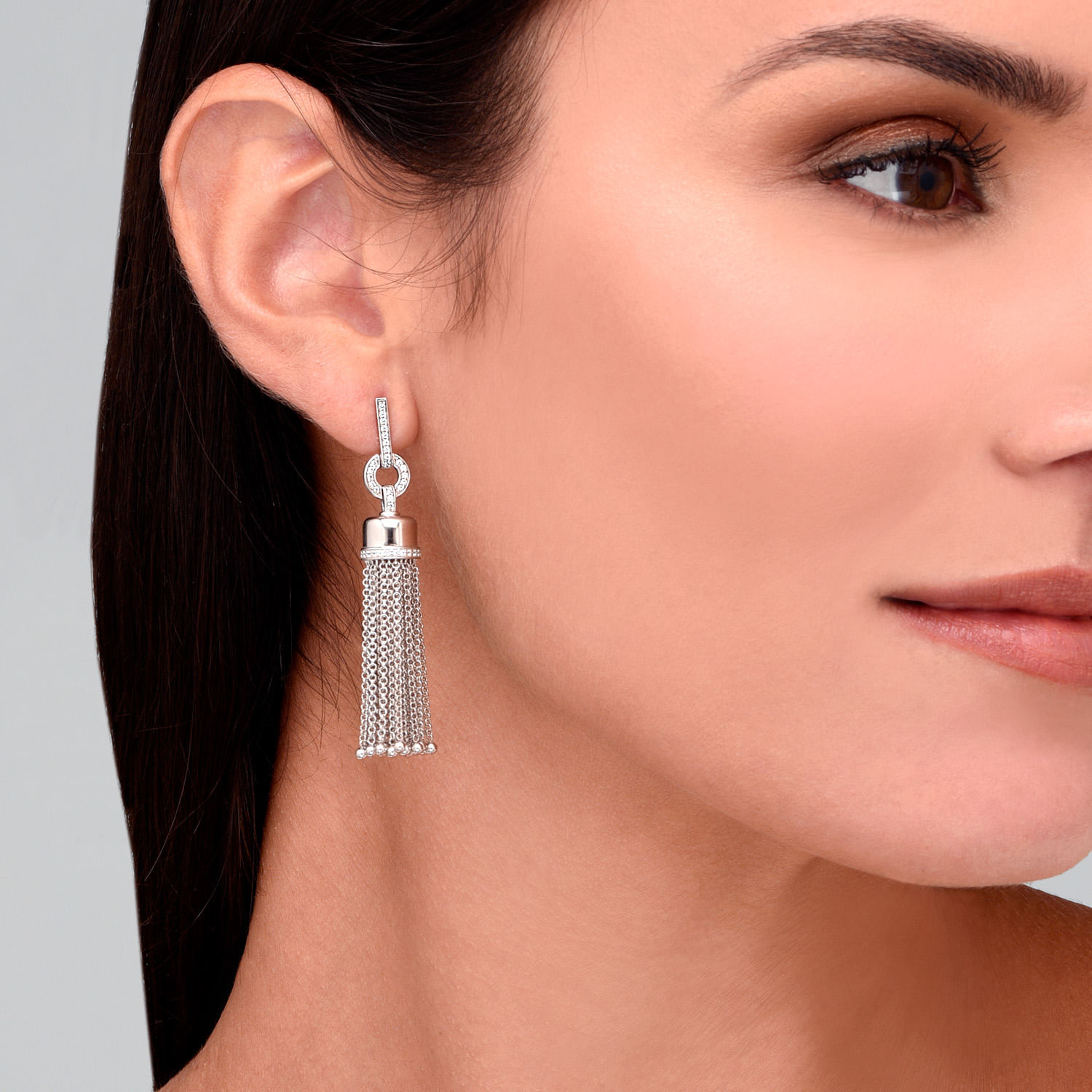 Brincos de argola com pendente de tassel de strass, em tom prateado, usados na orelha de mulher com pele clara e cabelo castanho escuro.