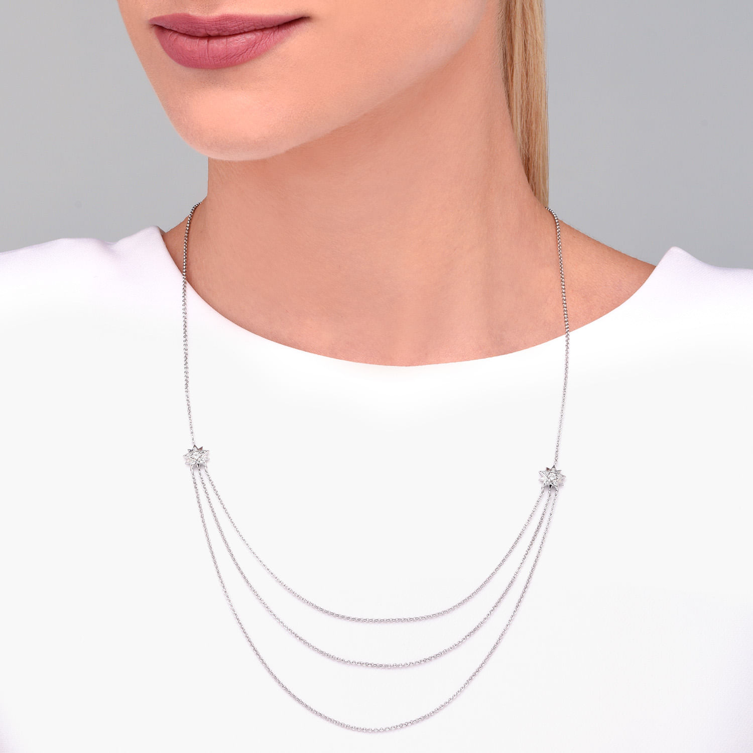 Colar prateado com três correntes e duas estrelas decorativas com pedras brilhantes, usado por uma mulher com blusa branca.