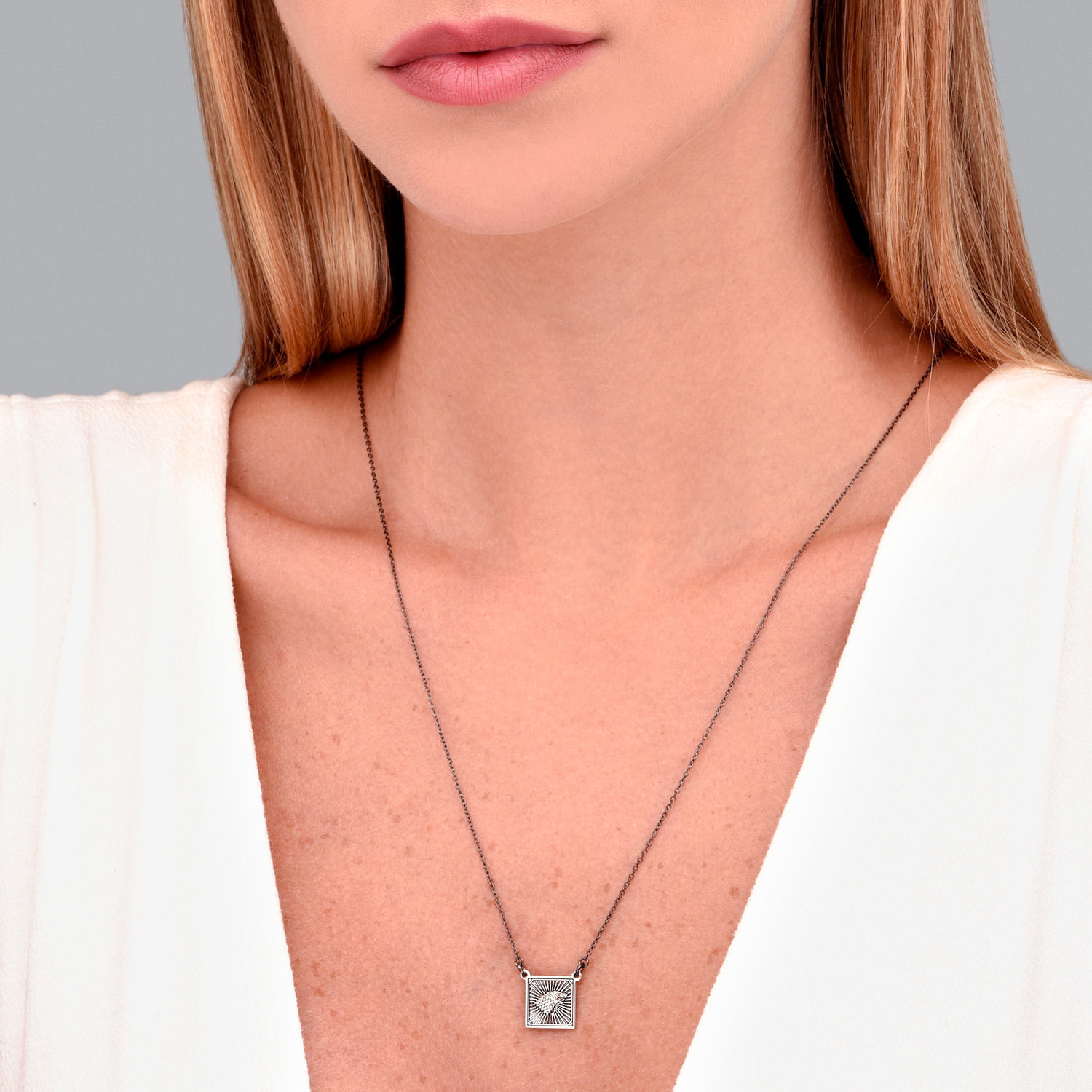 Colar com pingente quadrado prateado com detalhes decorativos, usado por uma mulher com pele clara, cabelo castanho claro e blusa branca.