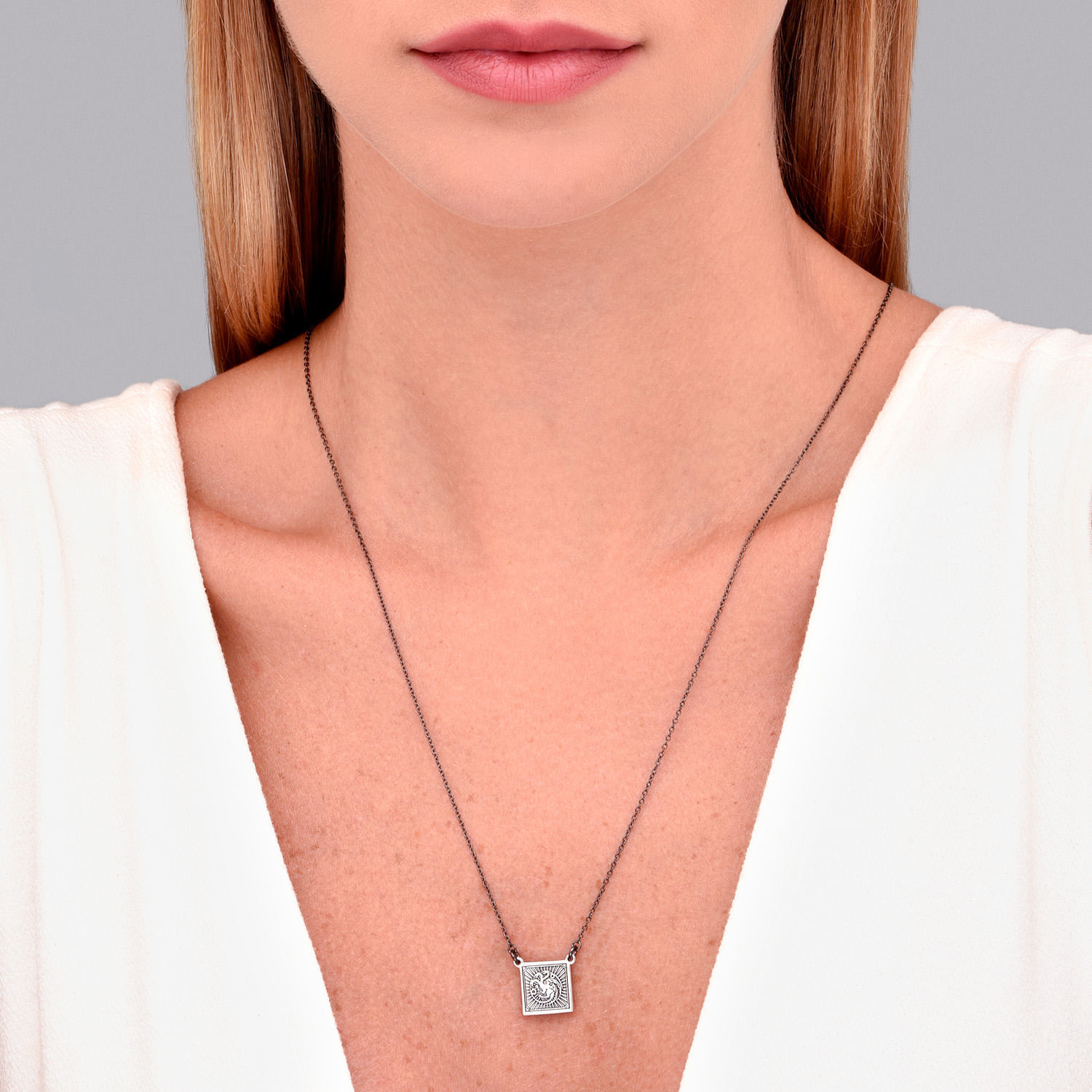 Mulher usando colar com pingente quadrado decorado, vestindo blusa branca de alças finas.