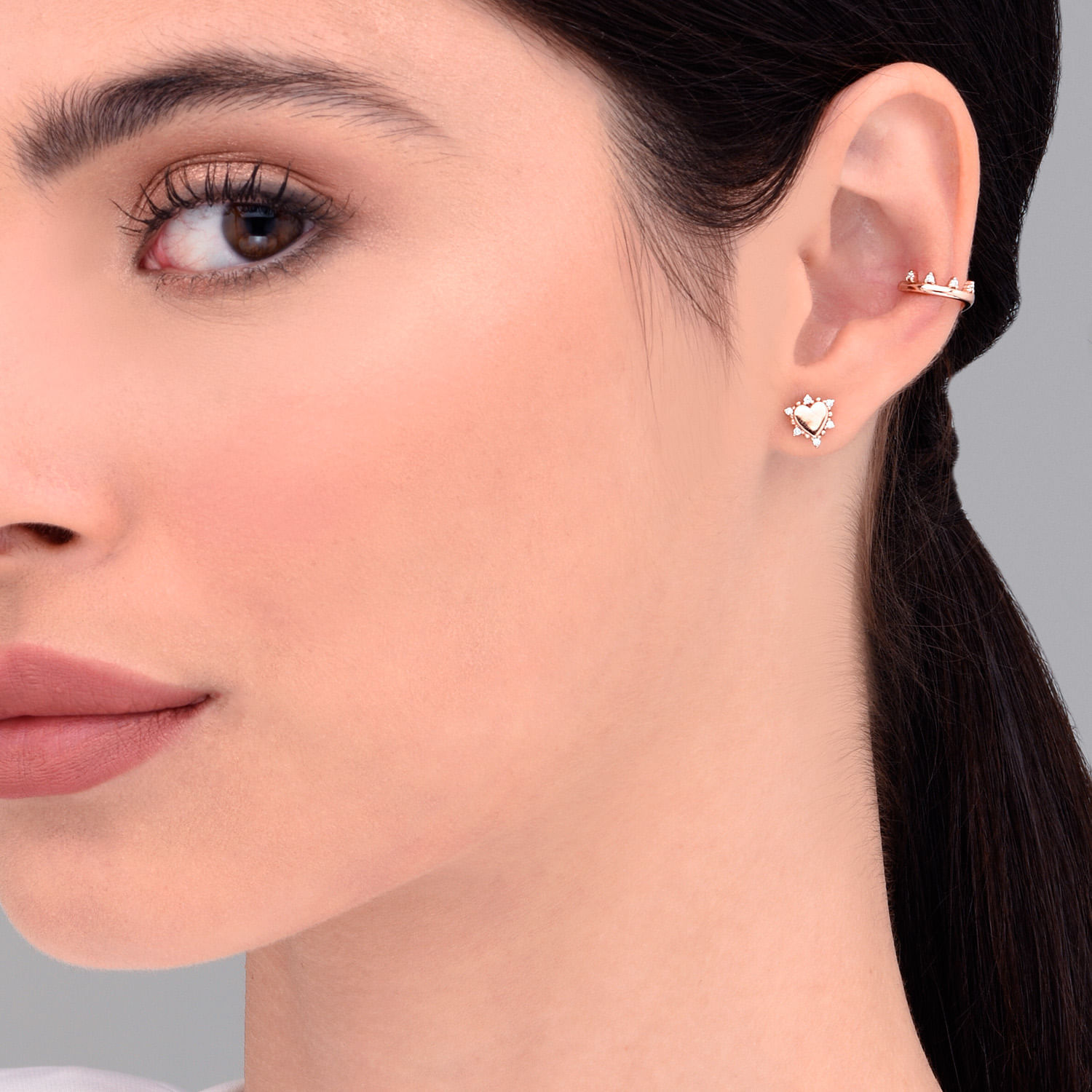 Close-up da orelha de uma mulher usando três brincos: uma argola no helix superior, uma borboleta de coração na orelha média e uma joia na orelha baixa.