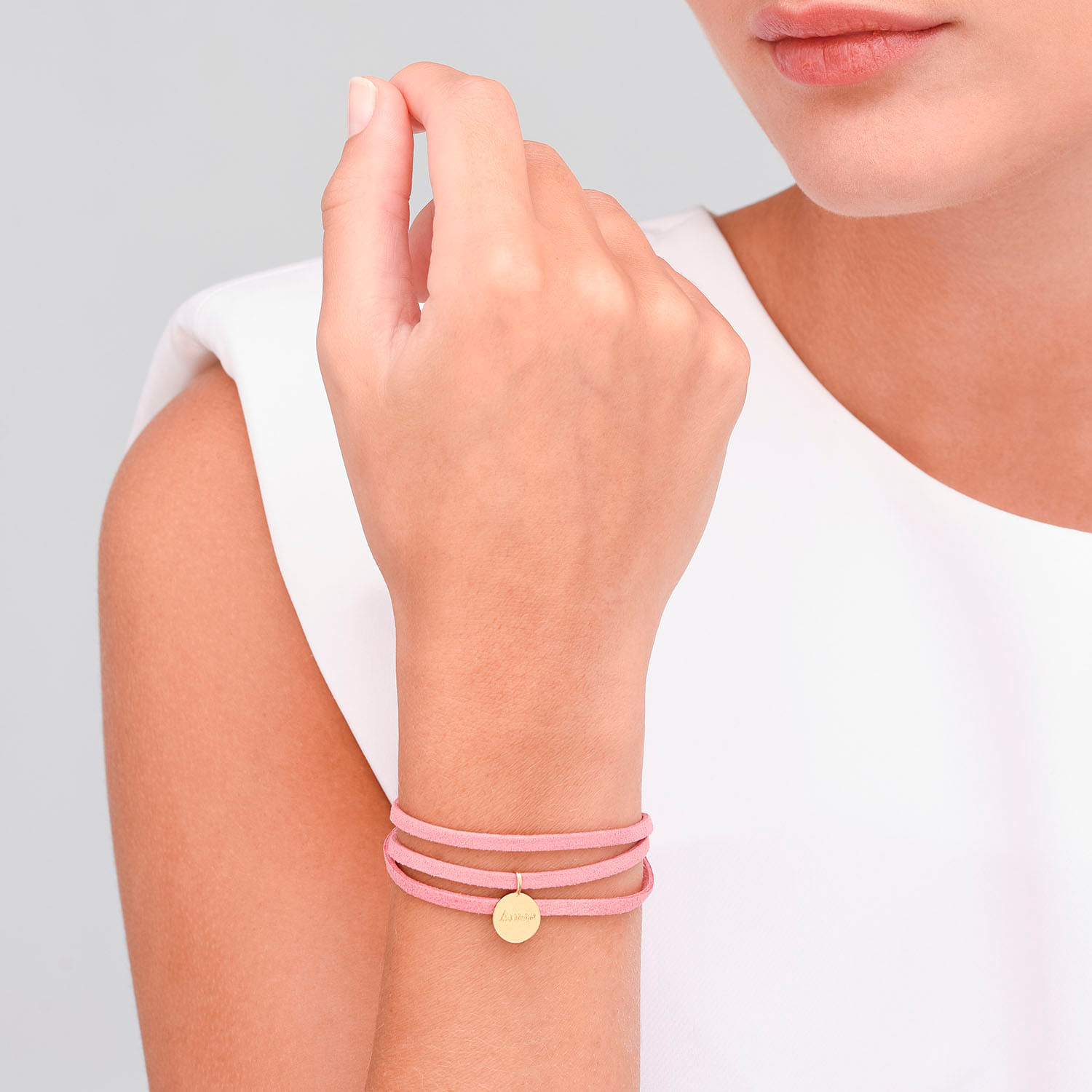 Mãos com três pulseiras rosadas e um pingente dourado, vestindo uma blusa branca, com fundo cinza claro.