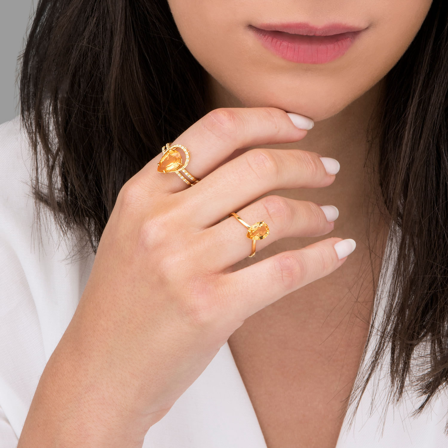 Mão de mulher com unhas brancas usando dois anéis dourados com pedras amarelas, uma no dedo médio e outra no indicador.
