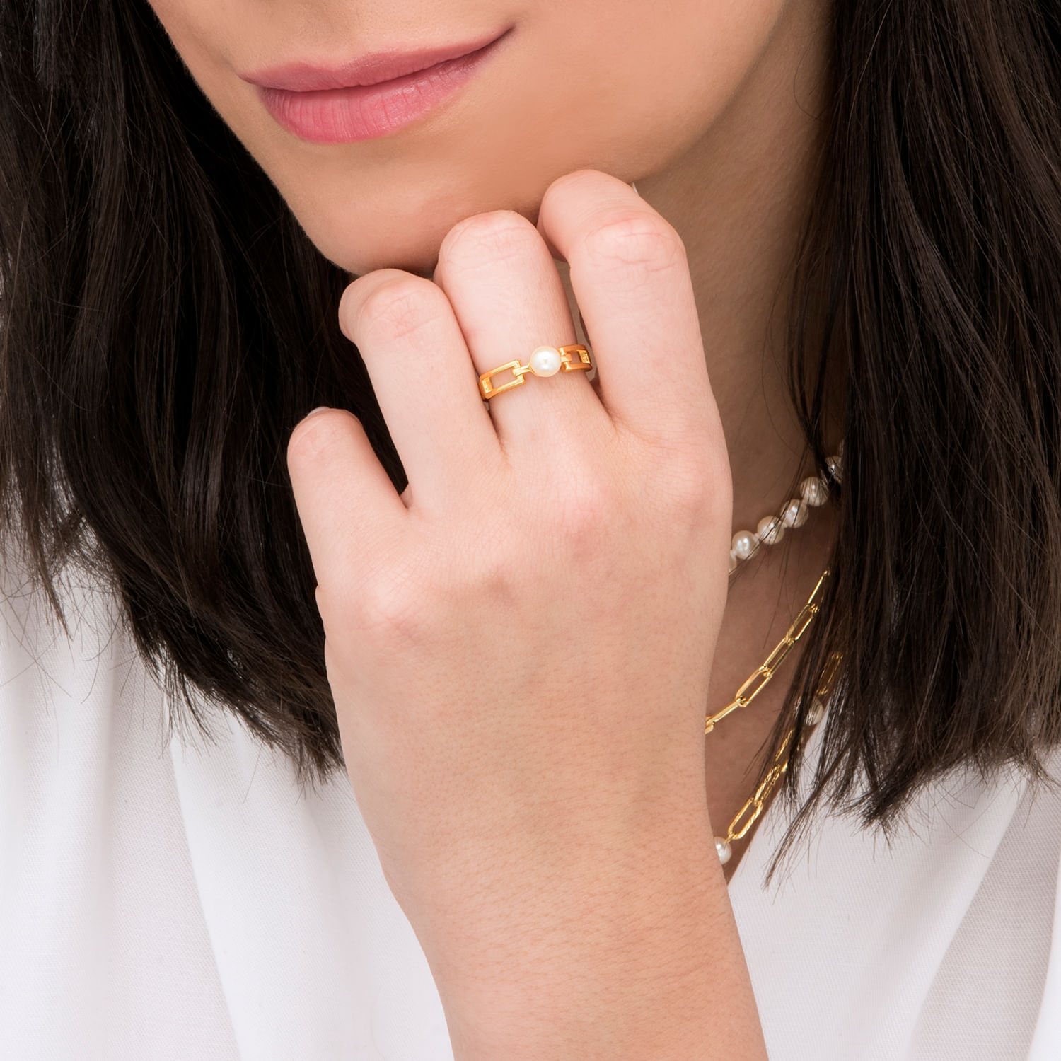 Close da mão e do pescoço de uma mulher com joias de ouro, colar de pérolas e um anel de ouro com uma pérola, perto dos lábios.