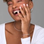Mulher sorrindo com cabelo cacheado, usando brincos, colar, pulseira e anel de joias brilhantes, com as mãos na face, vestindo blusa branca.