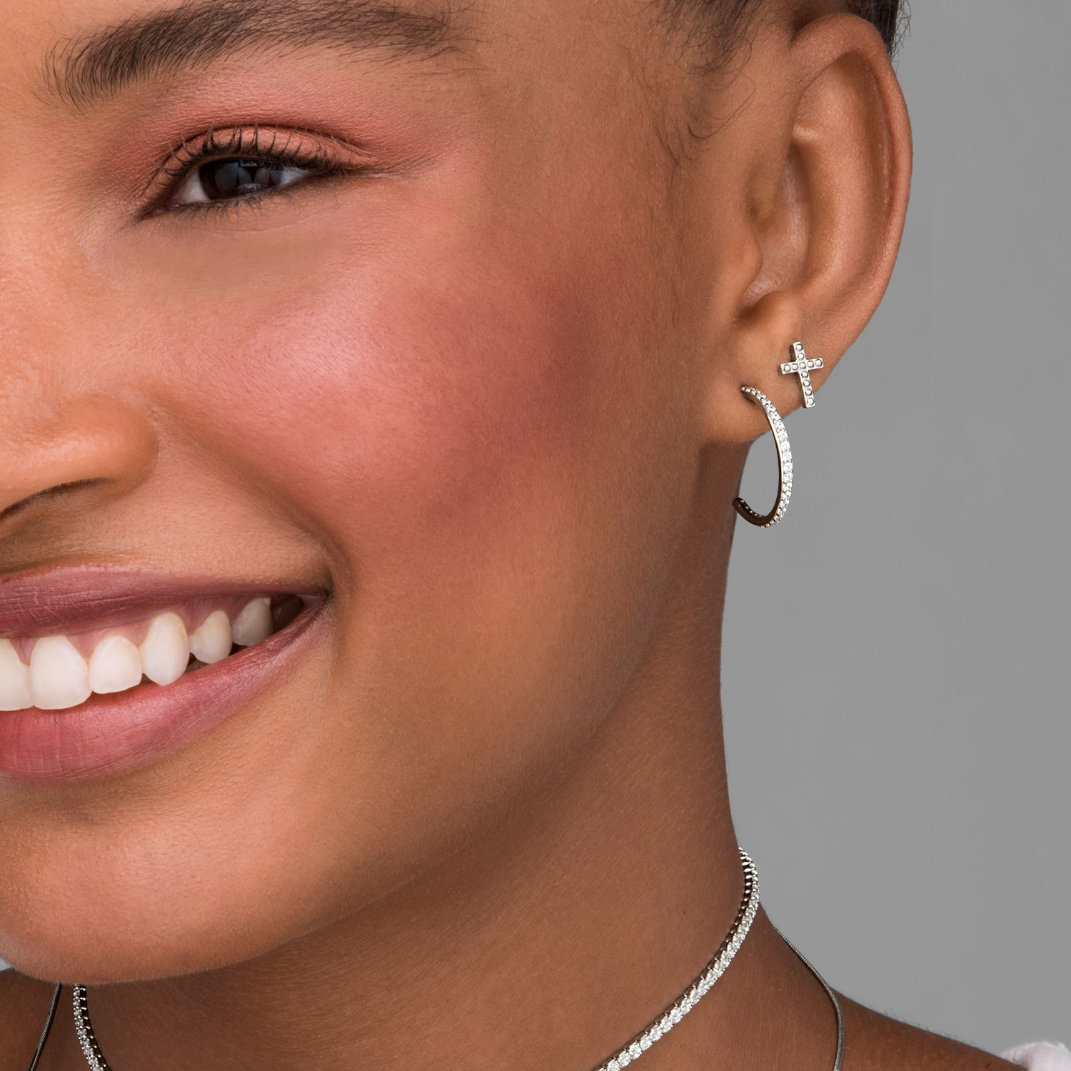 Close-up da metade do rosto de uma mulher sorridente, mostrando o olho, a boca, e um conjunto de brincos e colar com pedras brilhantes.