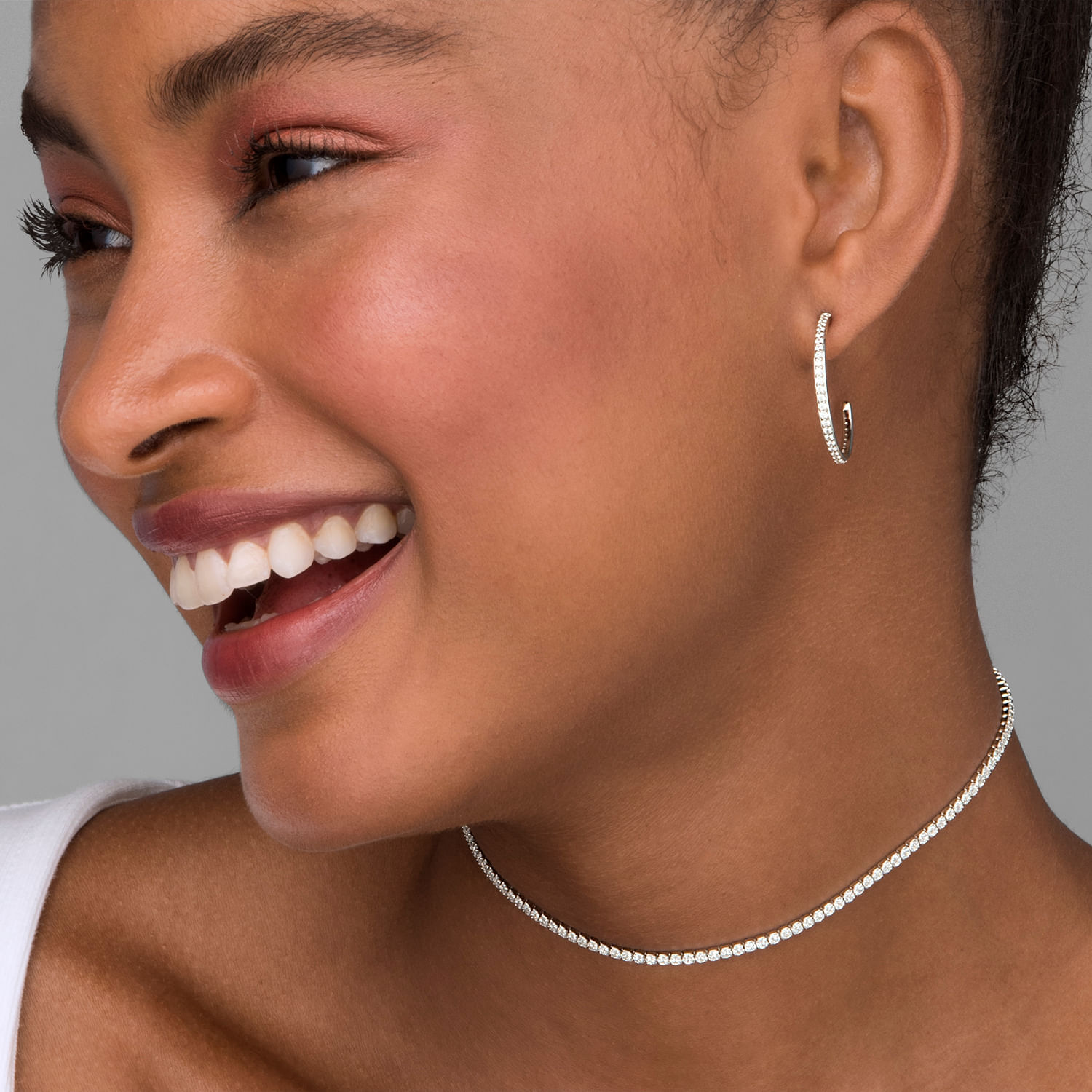 Close de uma mulher sorrindo, usando um brinco de argola de diamante, um colar de diamantes e uma blusa branca.