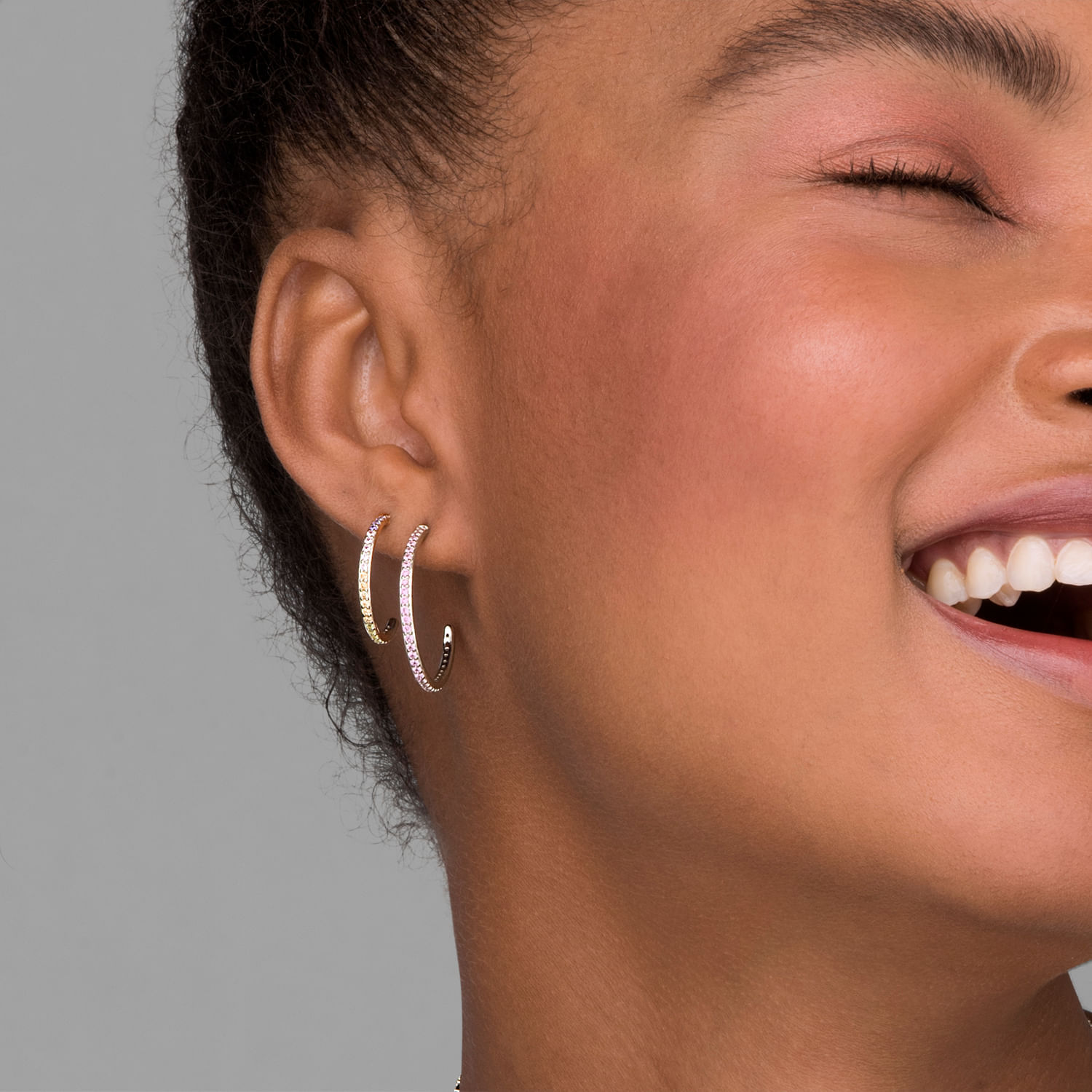 Mulher sorridente com brincos de argola dourados e prateados, usando maquiagem leve, com cabelo curto e cacheado.