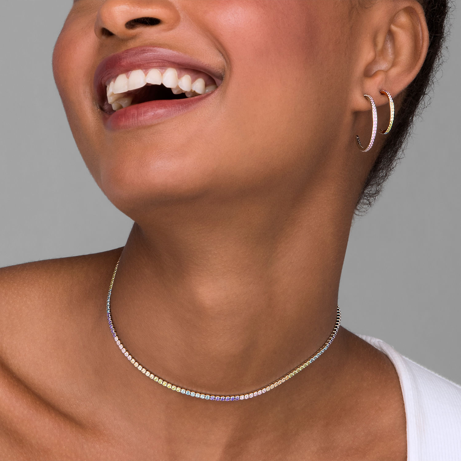 Pessoa sorrindo, usando brincos de argola e colar de pedras brilhantes, com pele morena, fundo cinza.