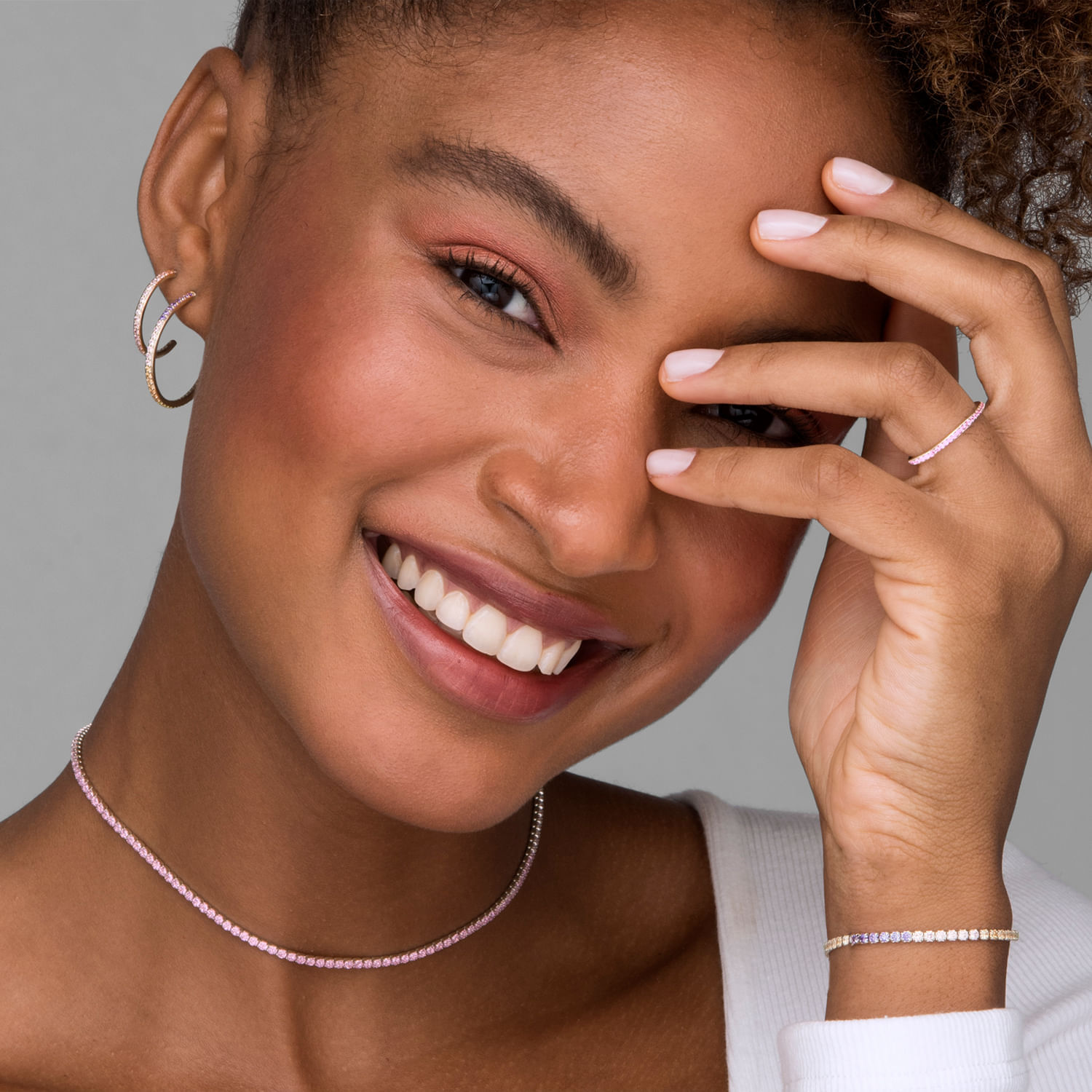 Mulher sorridente usando brincos, colar, pulseira e anel, com a mão na testa cobrindo parcialmente o rosto.