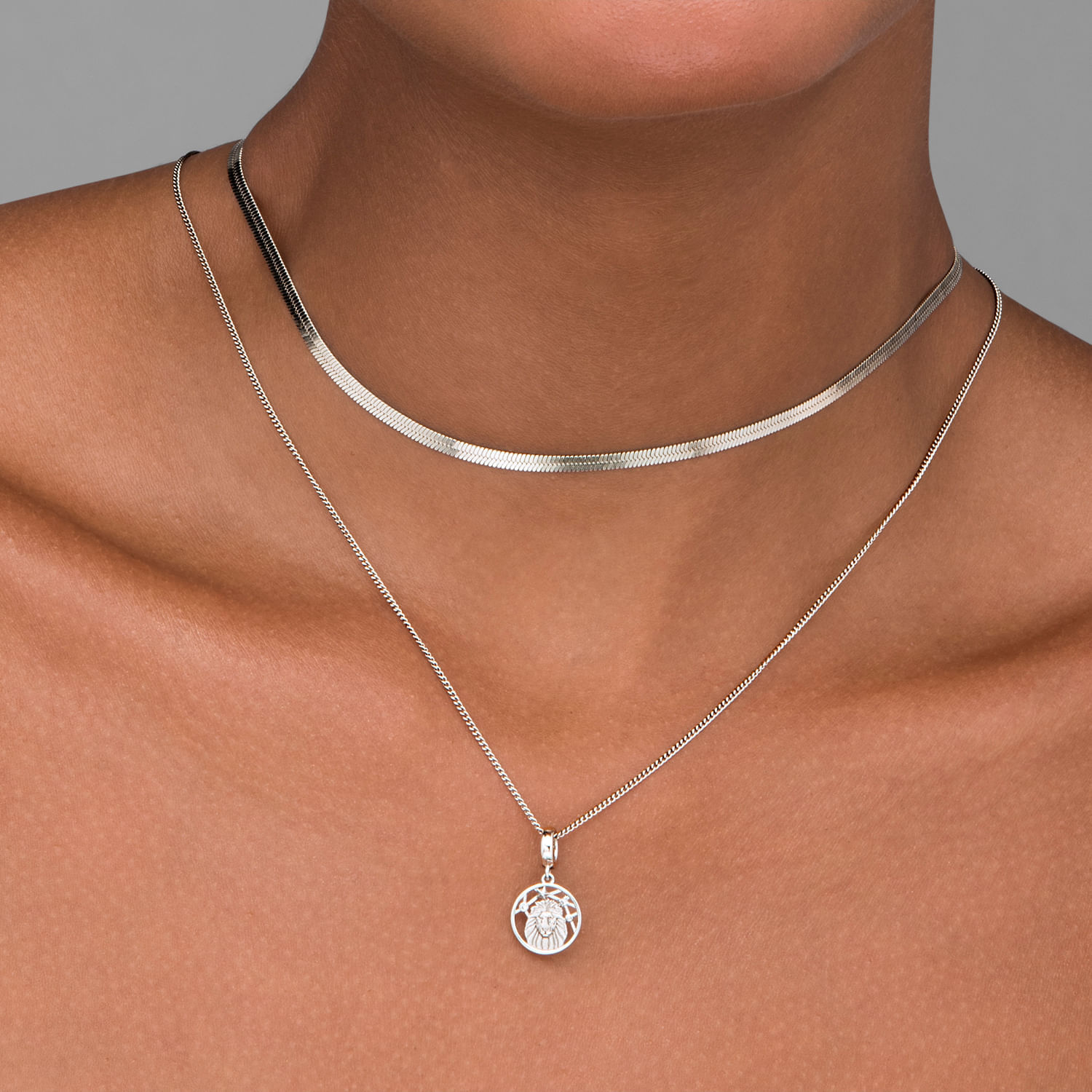 Colar e pingente prateados com dois colares finos, um com escudo e símbolo dentro, usados no pescoço de uma pessoa.