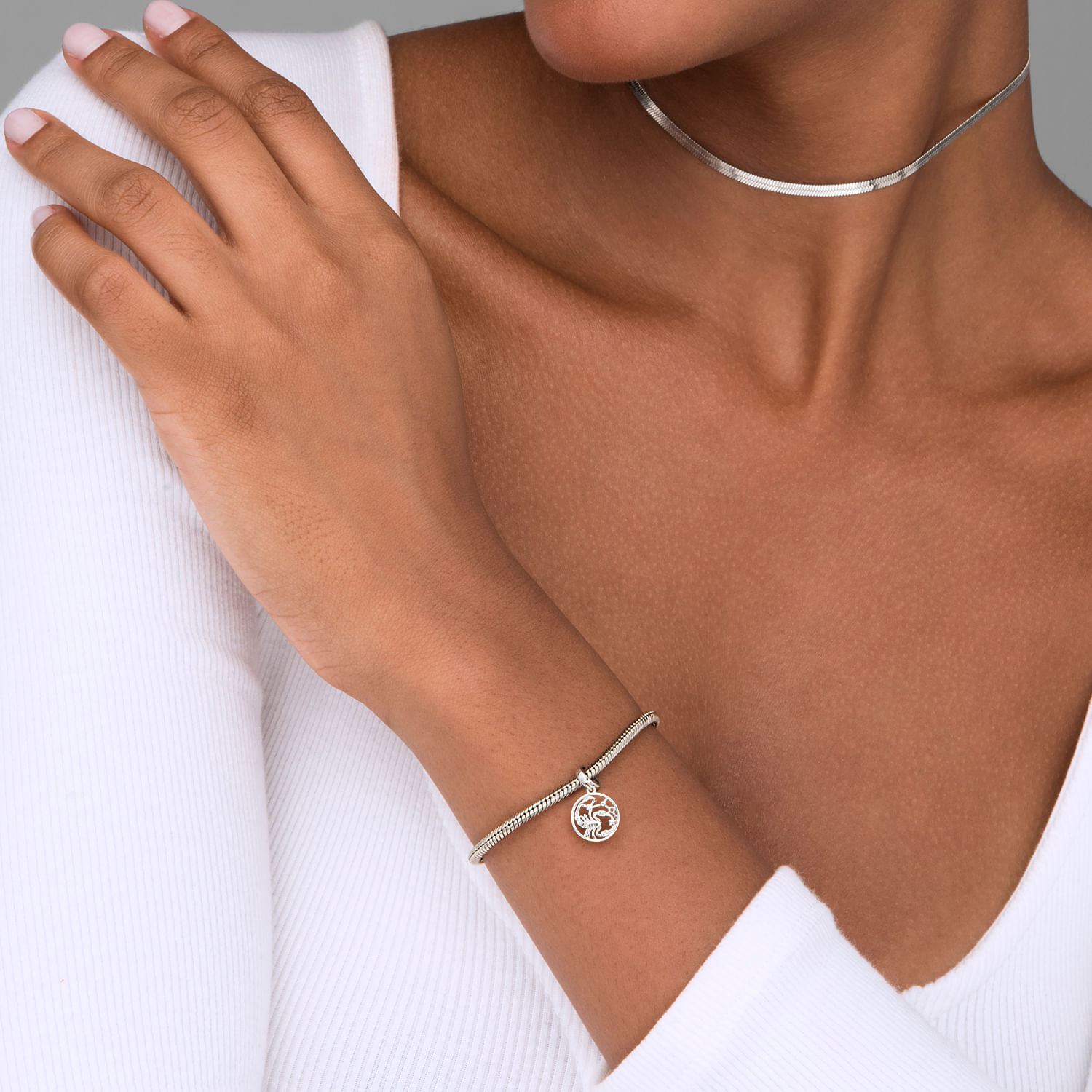 Mão de mulher usando uma pulseira prateada com pingente redondo e uma choker prateada. Ela está com uma blusa branca e a pele morena.