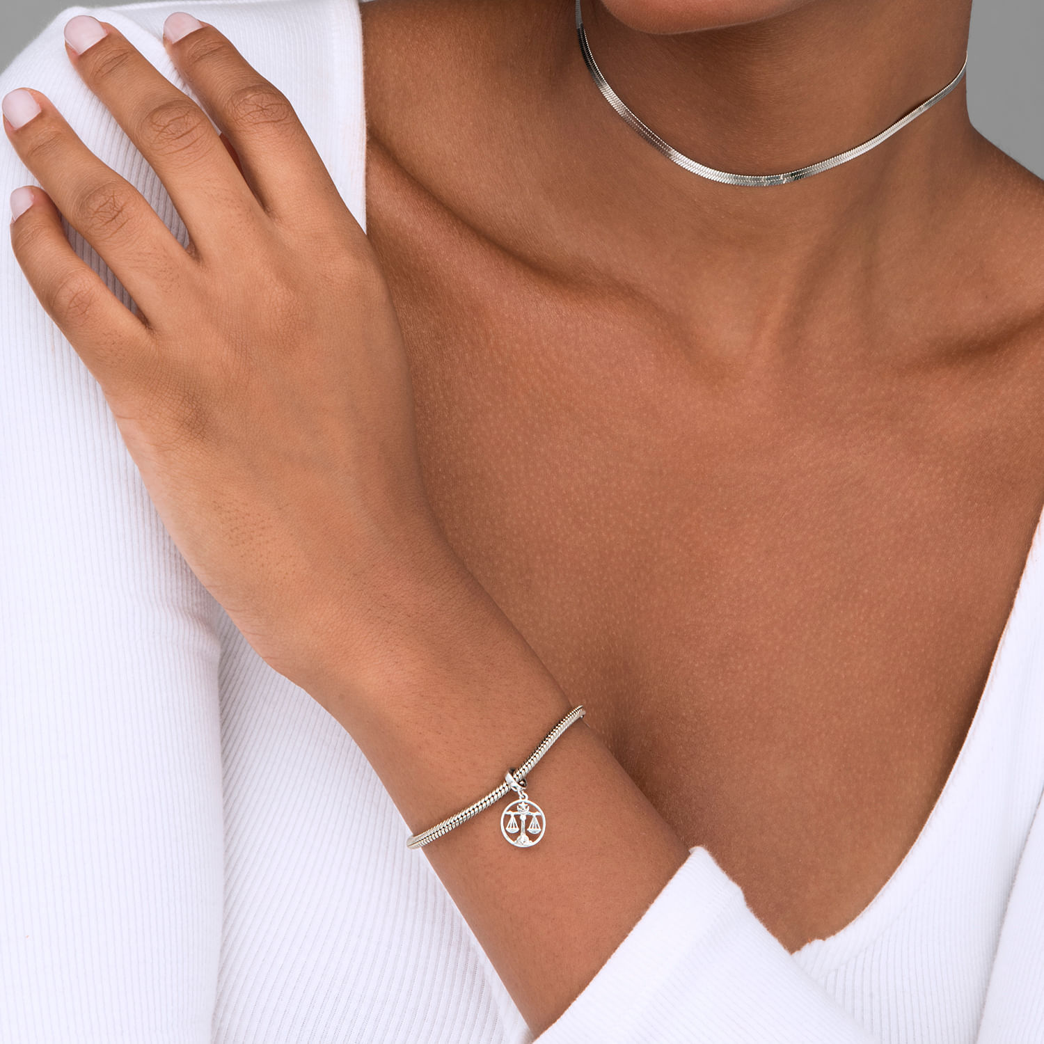 Mão de uma mulher com pele morena, usando uma pulseira de prata com pingente e colar fino de prata, vestindo blusa branca de manga longa.