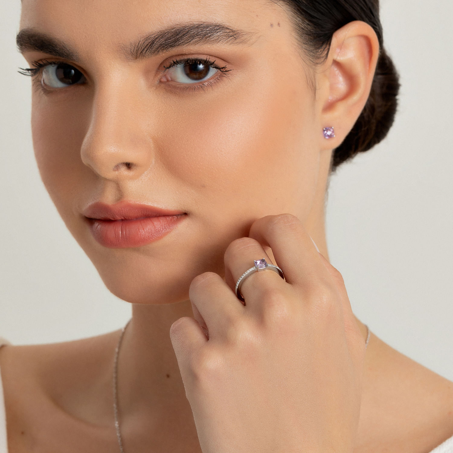 Close de uma mulher com olhos castanhos e pele clara usando brincos de pedras preciosas roxas e um anel com uma pedra preciosa roxa no dedo.