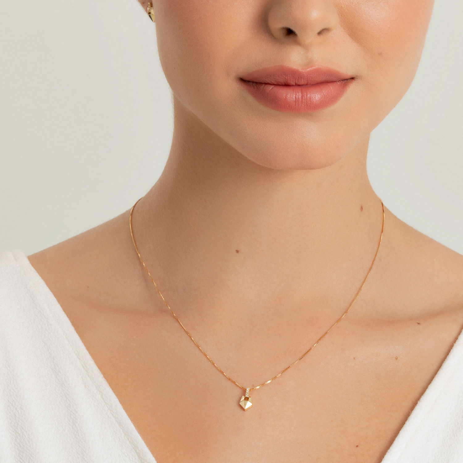 Colar delicado de corrente fina dourada com pingente hexagonal dourado na altura do pescoço de uma mulher.