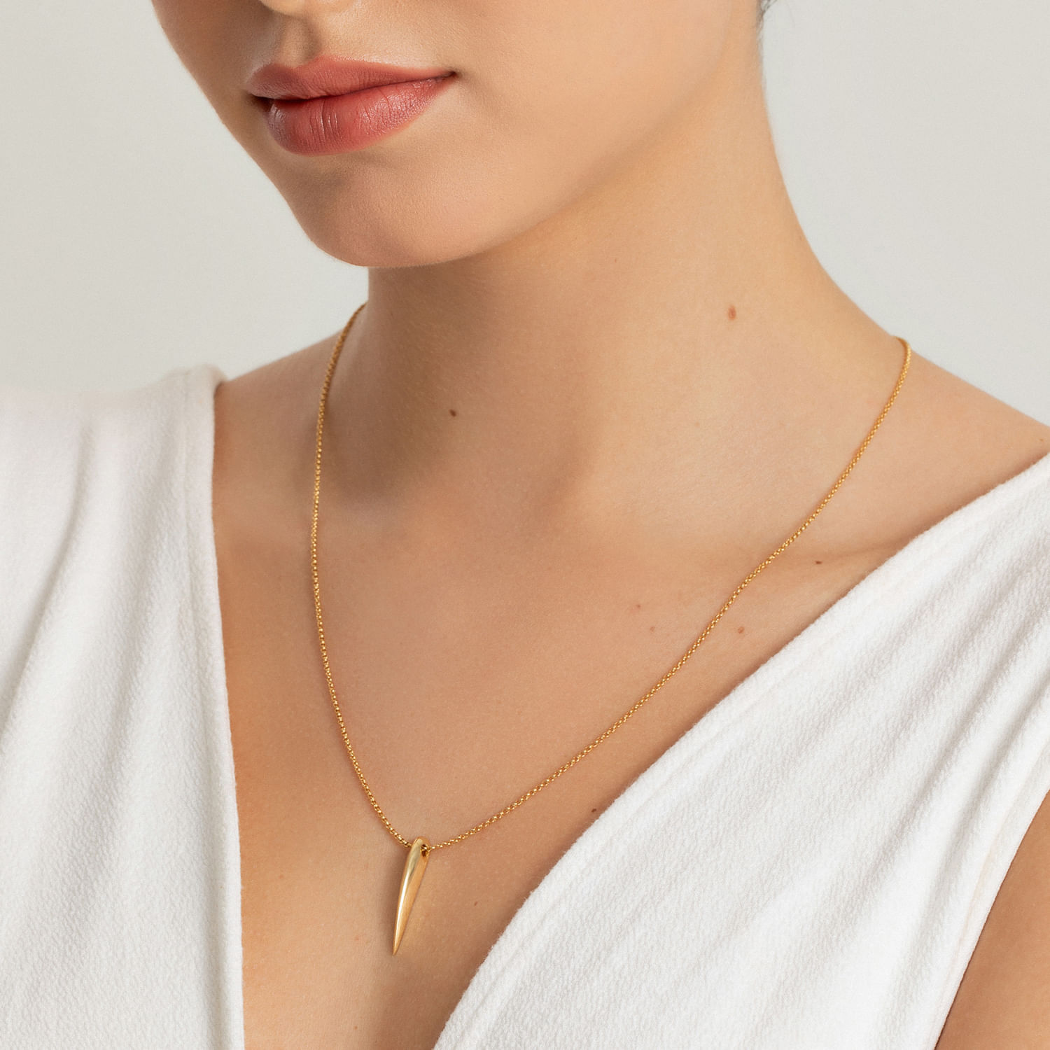 Colar dourado com pingente alongado em formato de lingueta, usado por uma mulher com roupa branca.
