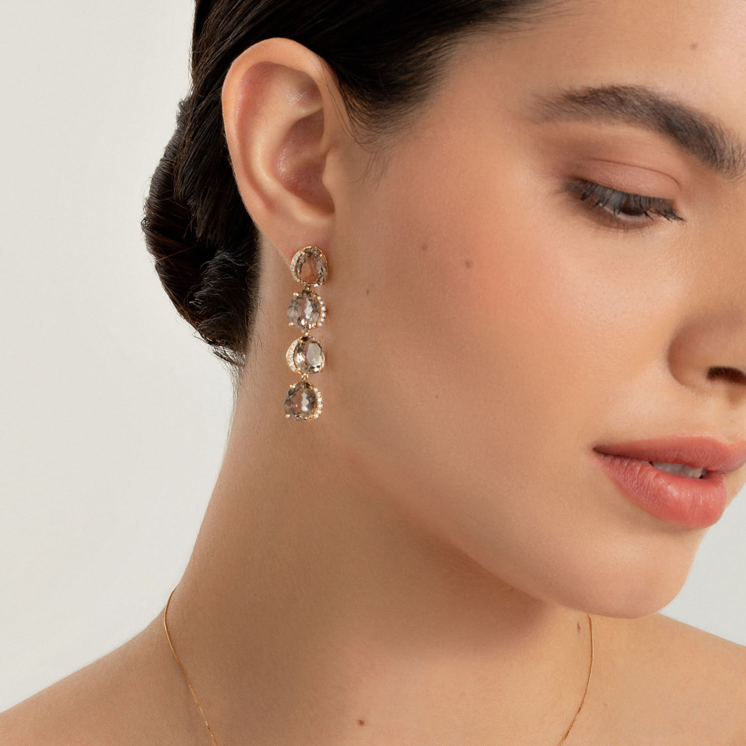 Close de uma mulher usando brincos pendurados com várias pedras e um delicado colar de ouro, com cabelo escuro e pele lisa.