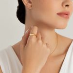 Mão de mulher usando anel dourado e colocando o dedo na garganta, com brincos e colar dourados, usando top branco.