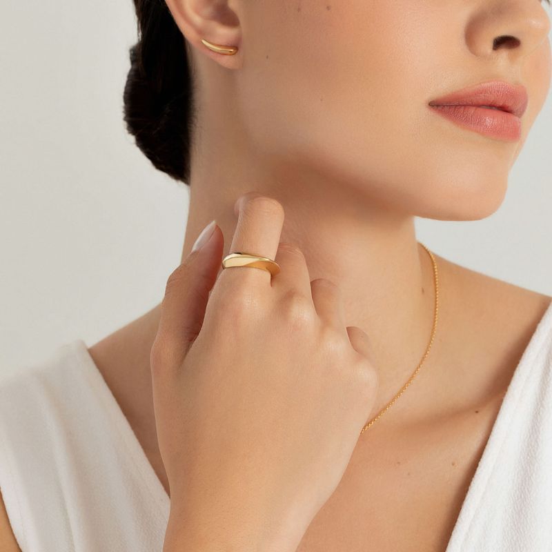 Mão de mulher usando anel dourado e colocando o dedo na garganta, com brincos e colar dourados, usando top branco.