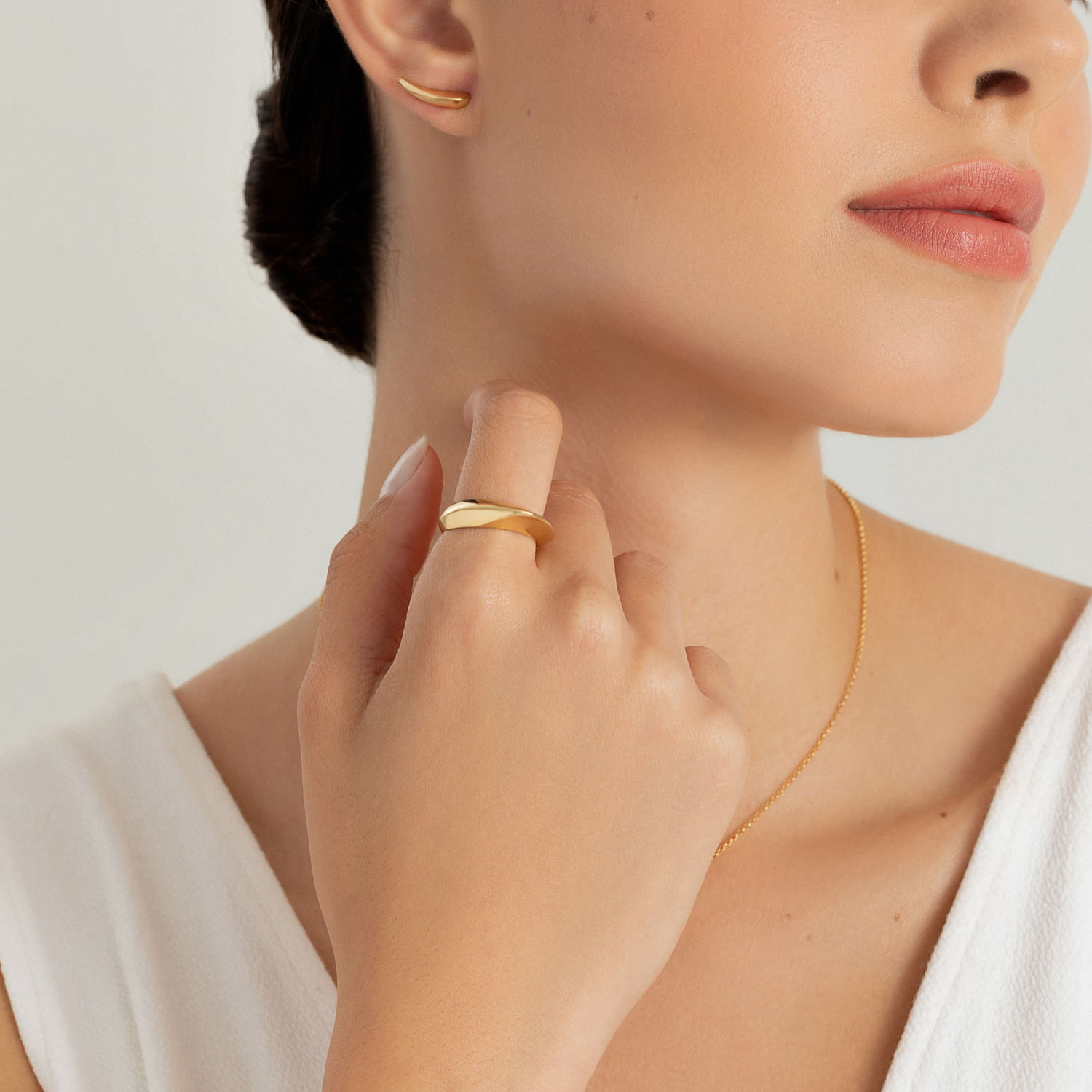 Mão de mulher usando anel dourado e colocando o dedo na garganta, com brincos e colar dourados, usando top branco.