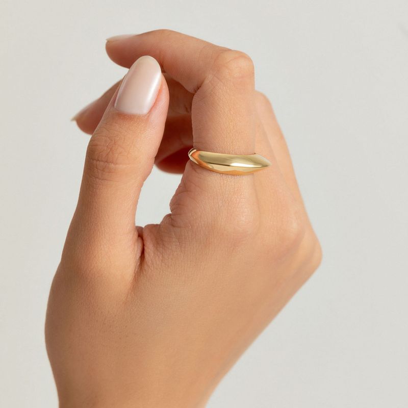 Mão com unha nude e anel dourado simples na ponta do dedo indicador, contra fundo branco.