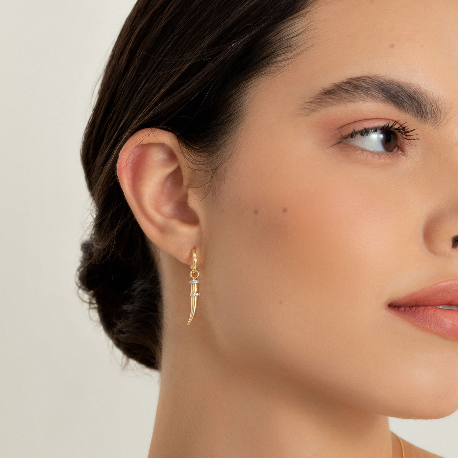 Close-up de uma mulher usando brinco dourado com pingente de dente e pequenos detalhes brilhantes na orelha esquerda.