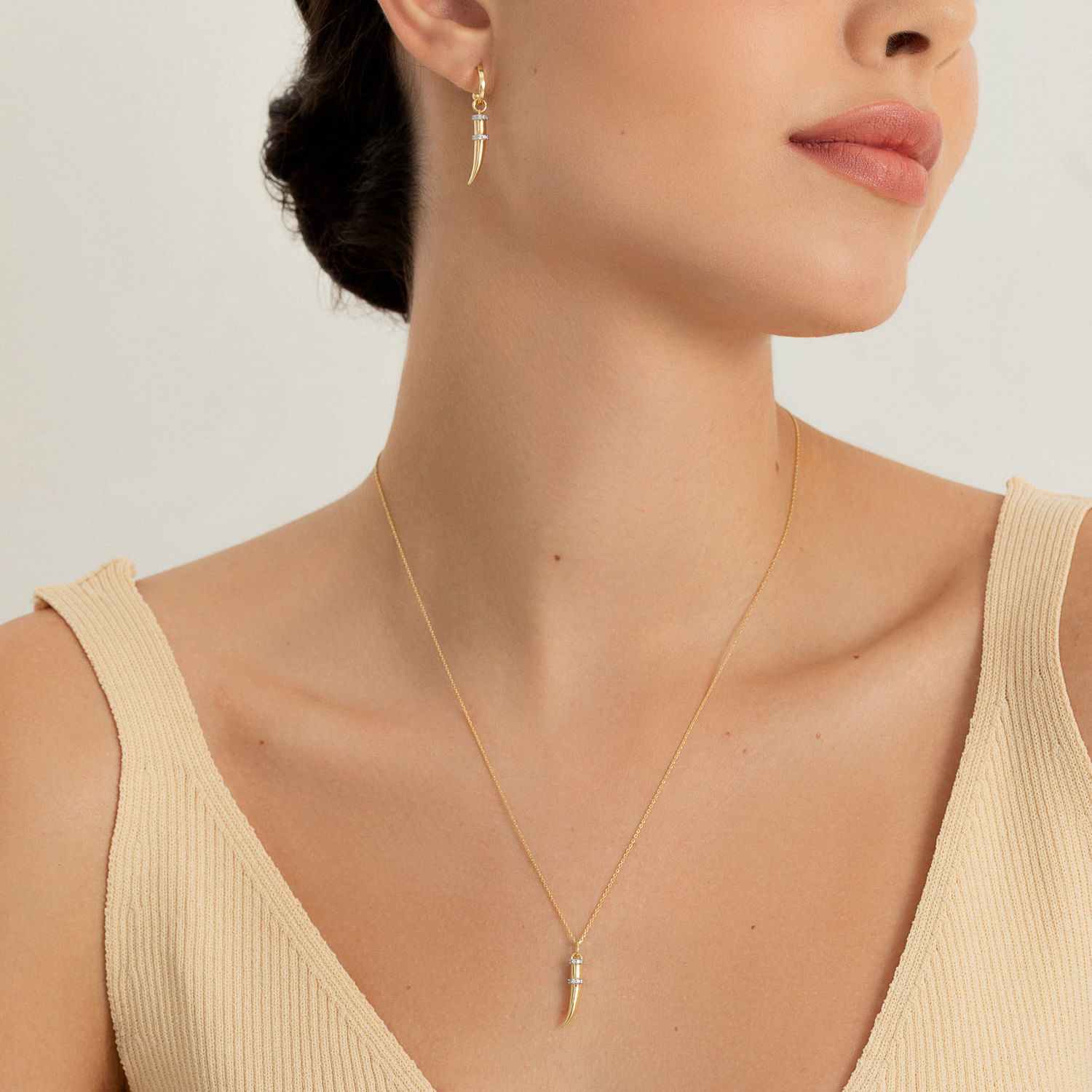 Mulher usando brincos de argola e colar dourados com pingente em forma de dente, vestindo top bege de alças finas.