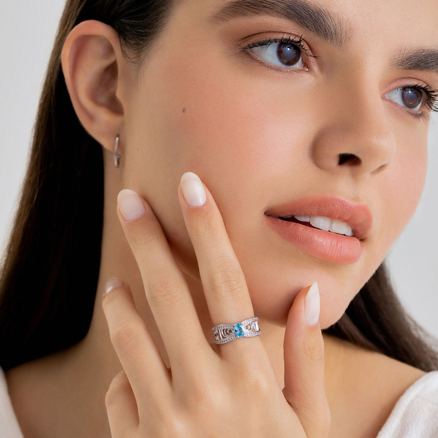 Close-up de uma mulher com pele clara, usando um anel com pedra azul na mão esquerda, e brincos de argola pequenos.