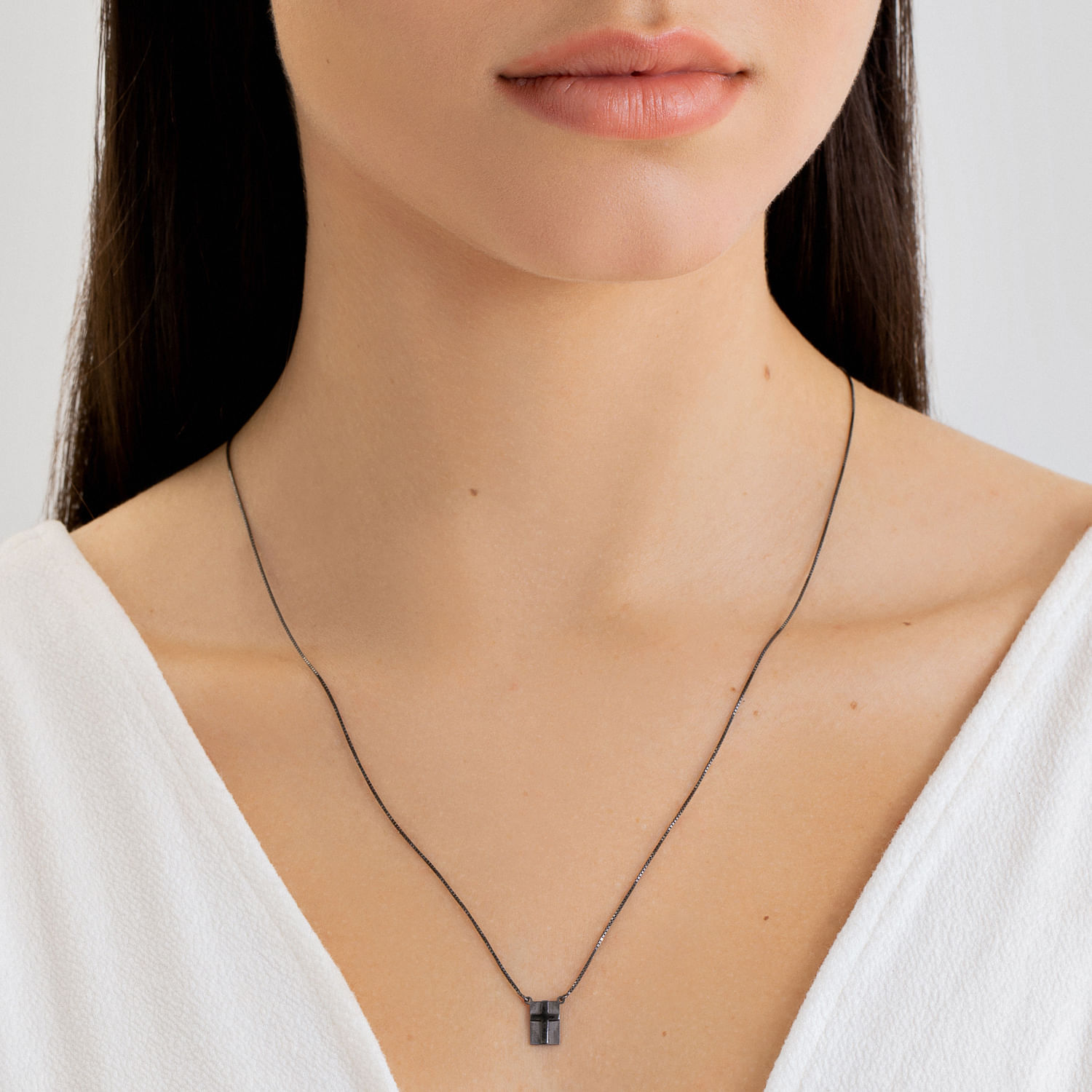 Colar fino de corrente preta com pingente quadrado preto, usado por uma mulher com blusa branca.