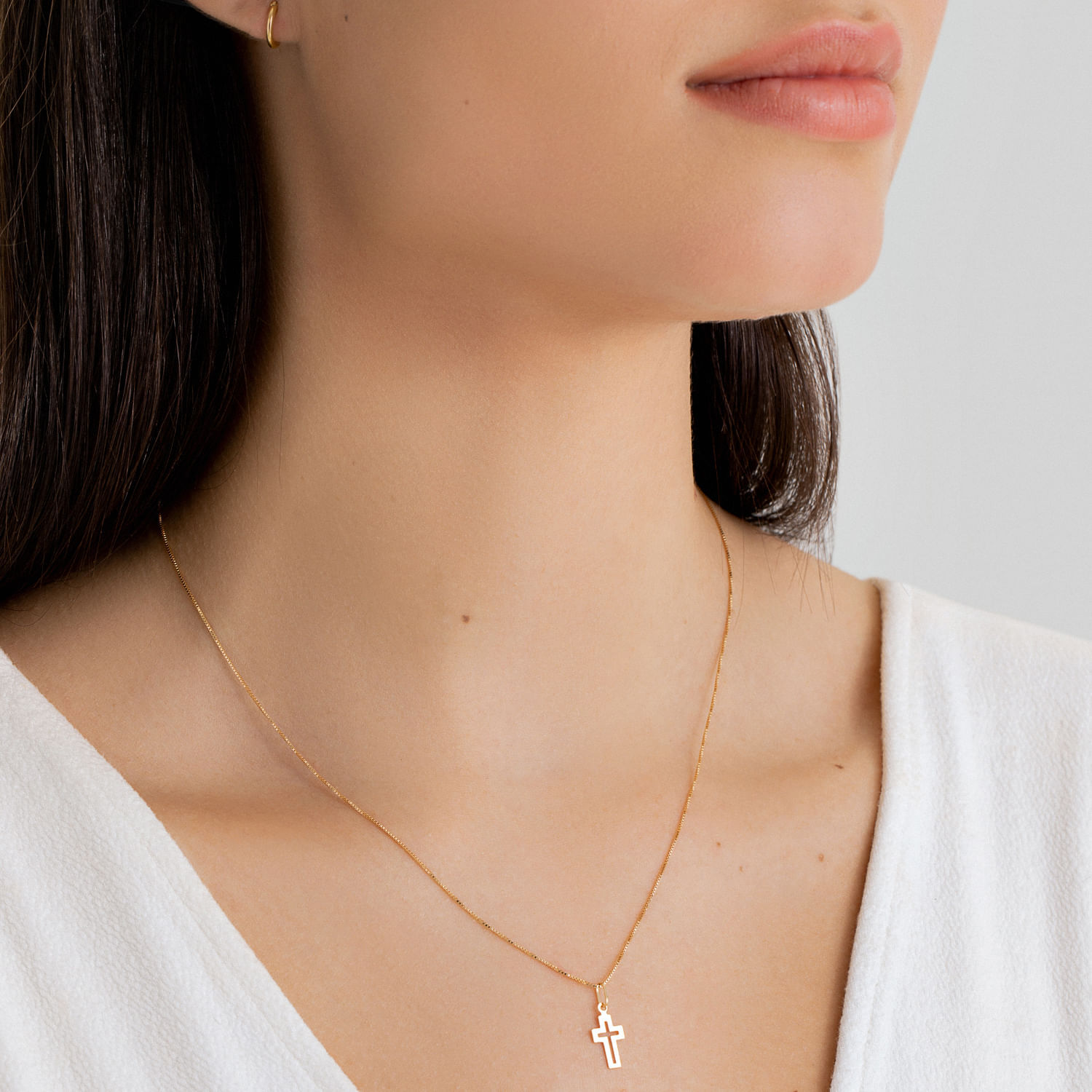 Colar delicado de corrente fina com pingente de cruz dourado, usado por uma mulher com pele clara e roupas brancas.