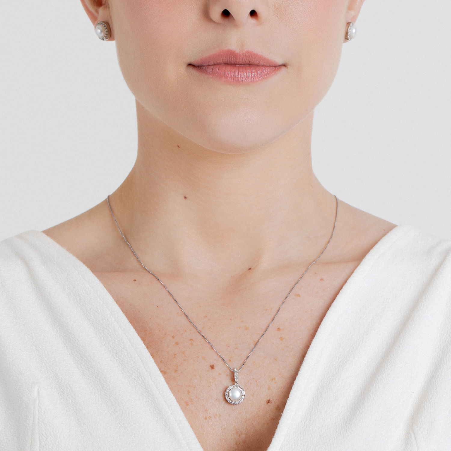 Colar delicado com pingente de pérola envolto por pequenos diamantes, usado por mulher com blusa branca.