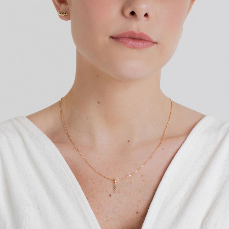 Mulher com colar e pingente de ouro com pedras verdes, vestida com blusa branca e acessórios de orelha.