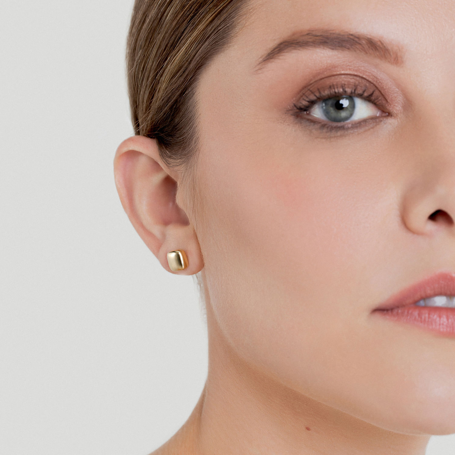 Close-up de uma mulher com pele clara, cabelo castanho e usando brincos dourados quadrados.
