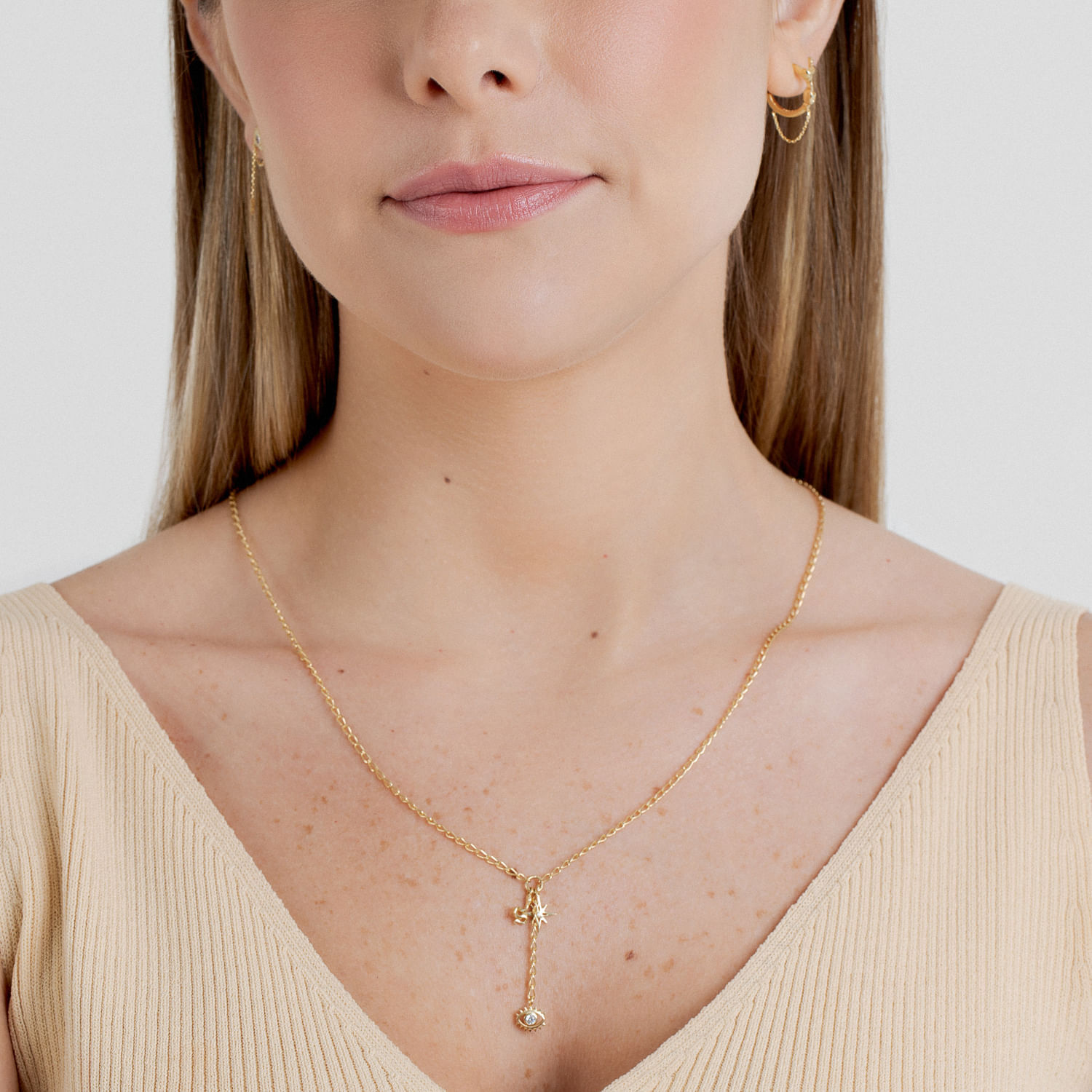 Colar dourado com pingente de estrela e uma pedra, usado por uma mulher com roupas de tom bege, com cabelo castanho claro e brincos dourados.