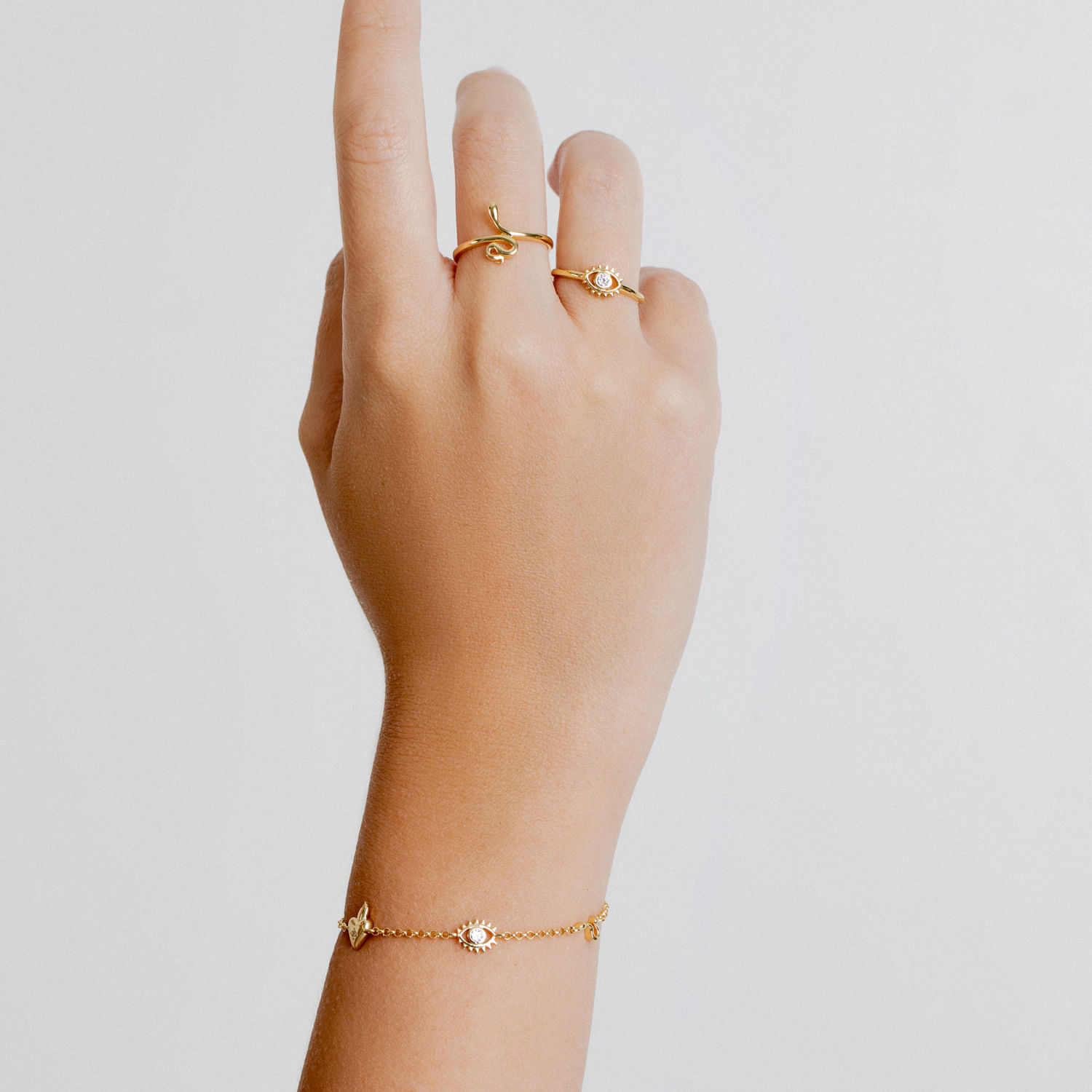 Mão com dois anéis dourados e uma pulseira dourada com pequenos pingentes, contra fundo branco.