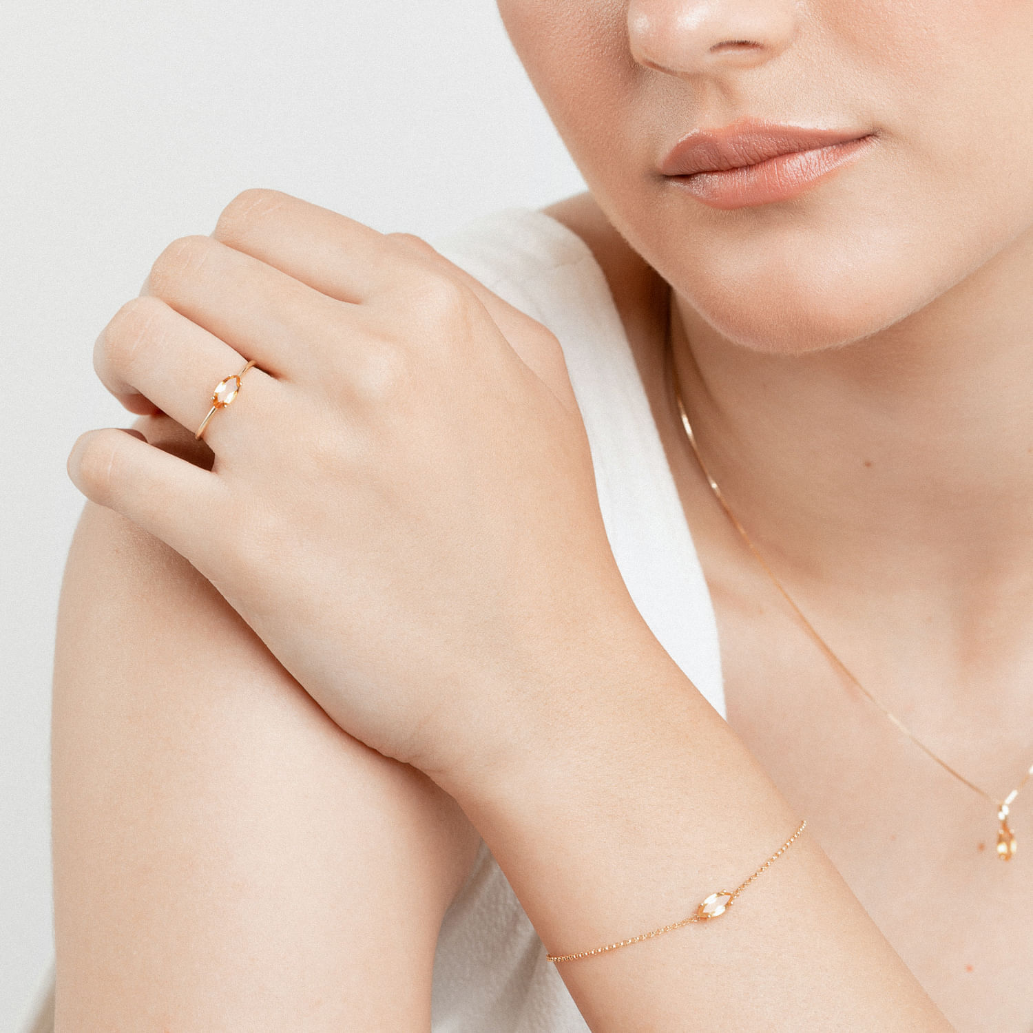 Mulher usando joias douradas delicadas, incluindo anel, pulseira e colar, com destaque para uma pedra branca no centro.