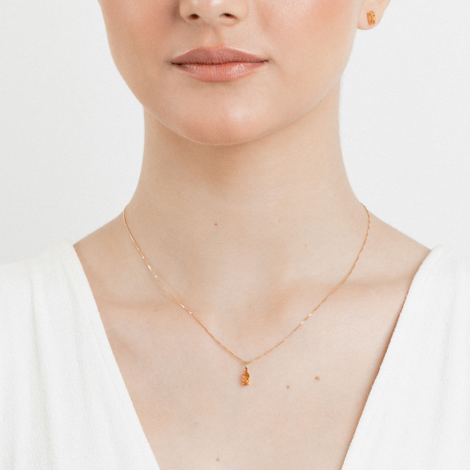 Colar delicado dourado com pingente de pedra laranja, usado por uma mulher de pele clara e camisa branca.