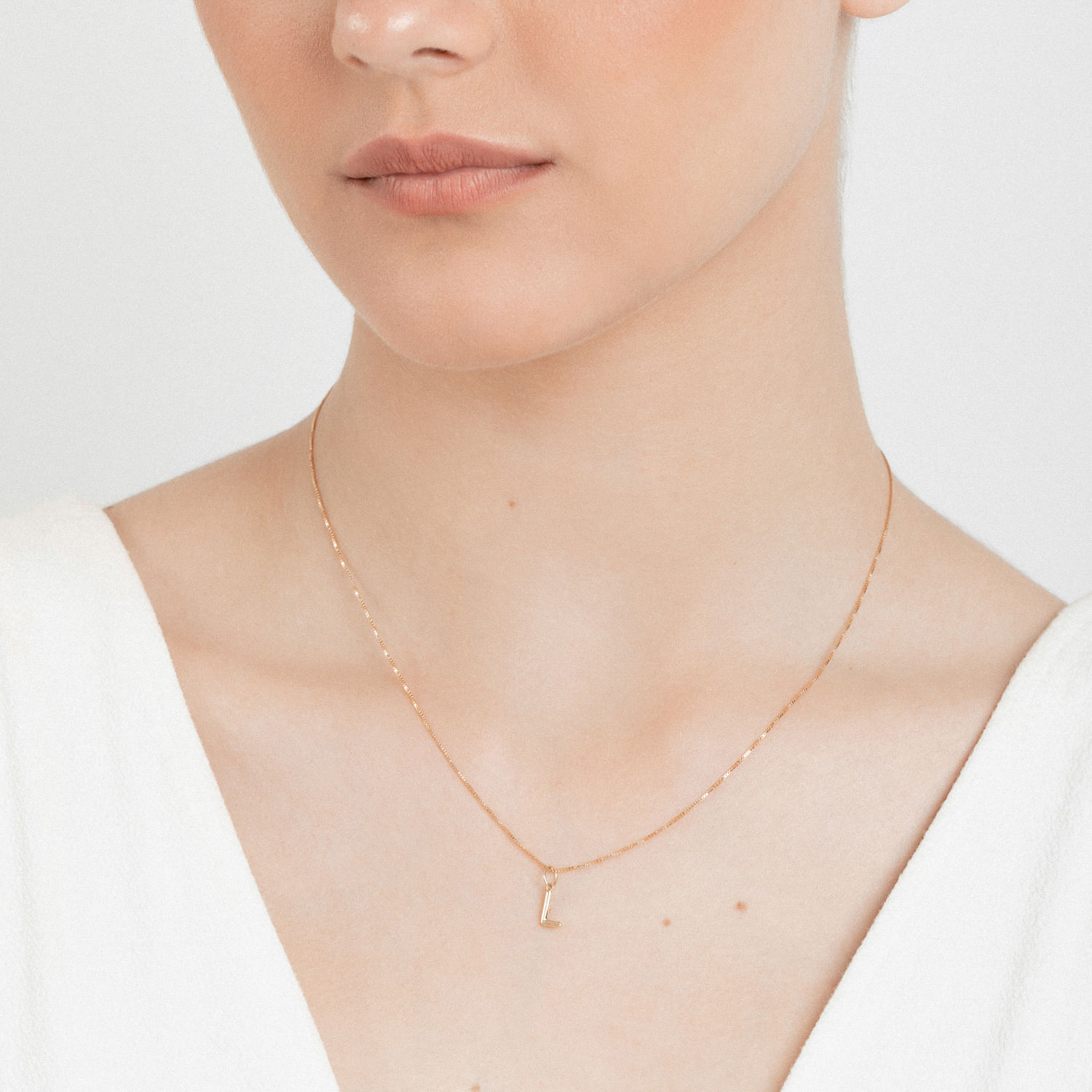 Colar fino de corrente delicada com pingente em forma de letra "L" na cor dourada, usado por uma mulher com pele clara e roupa branca.