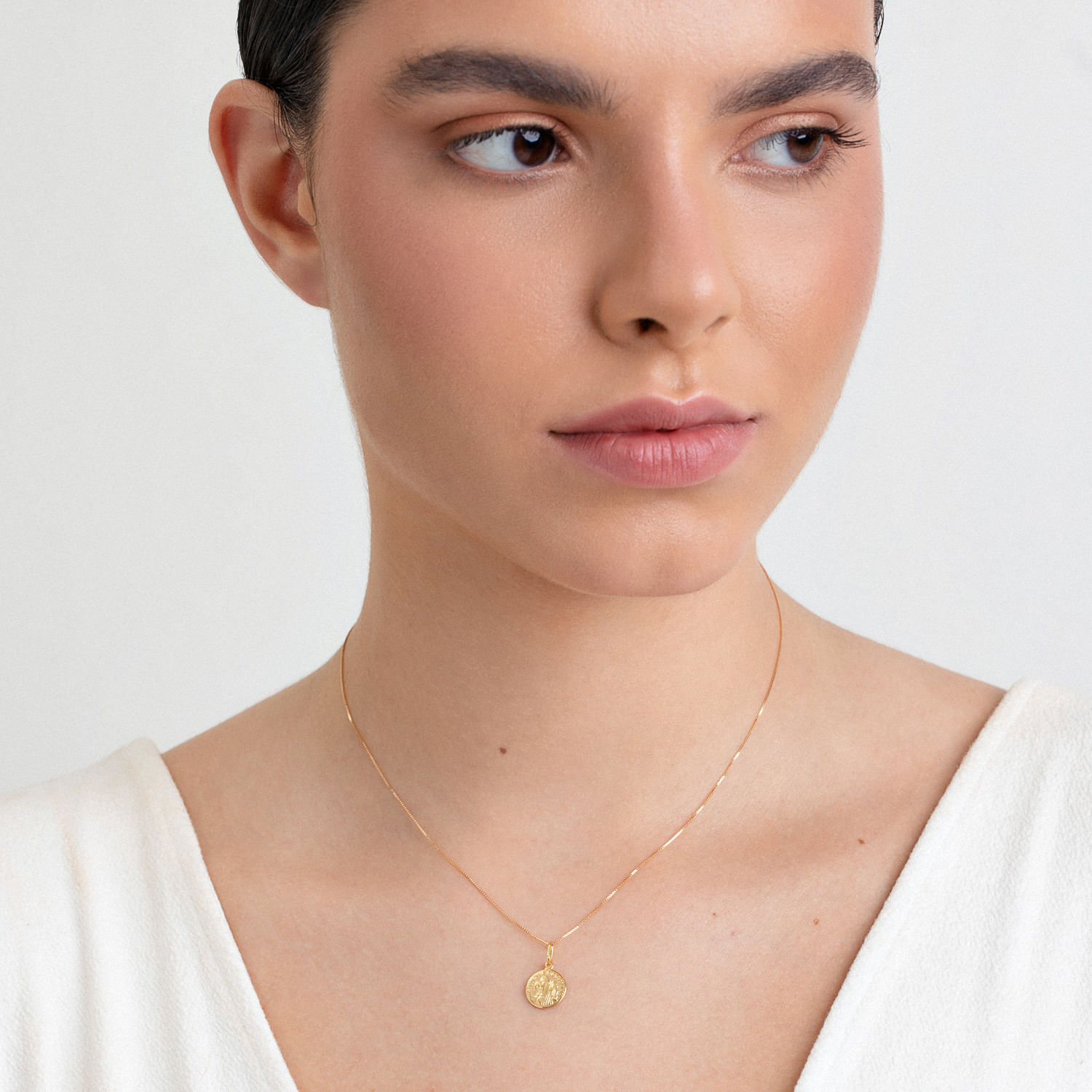 Mulher com cabelo escuro usando colar delicado com pingente dourado e blusa branca.