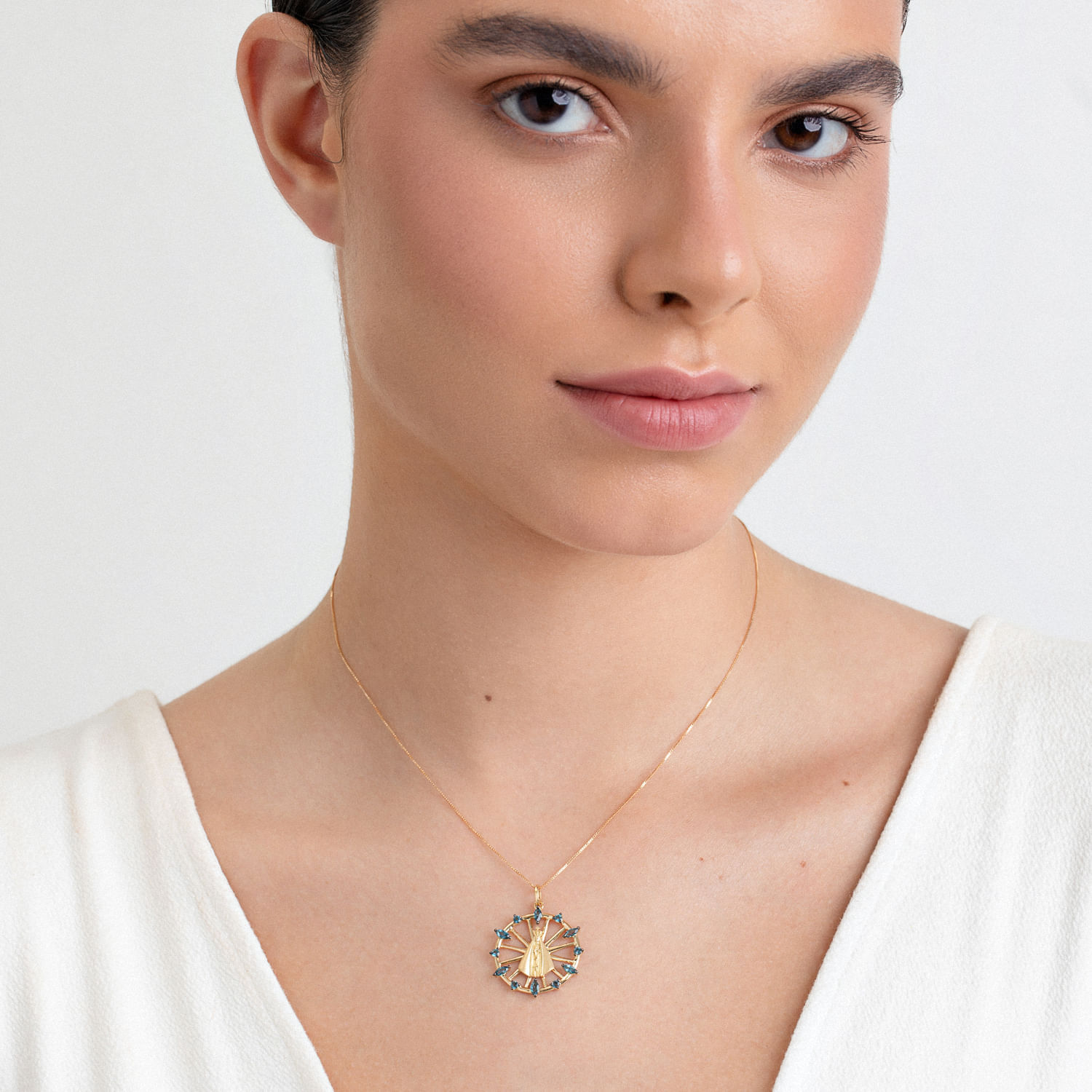 Mulher usando colar dourado com pingente circular com detalhes coloridos, vestindo blusa branca.