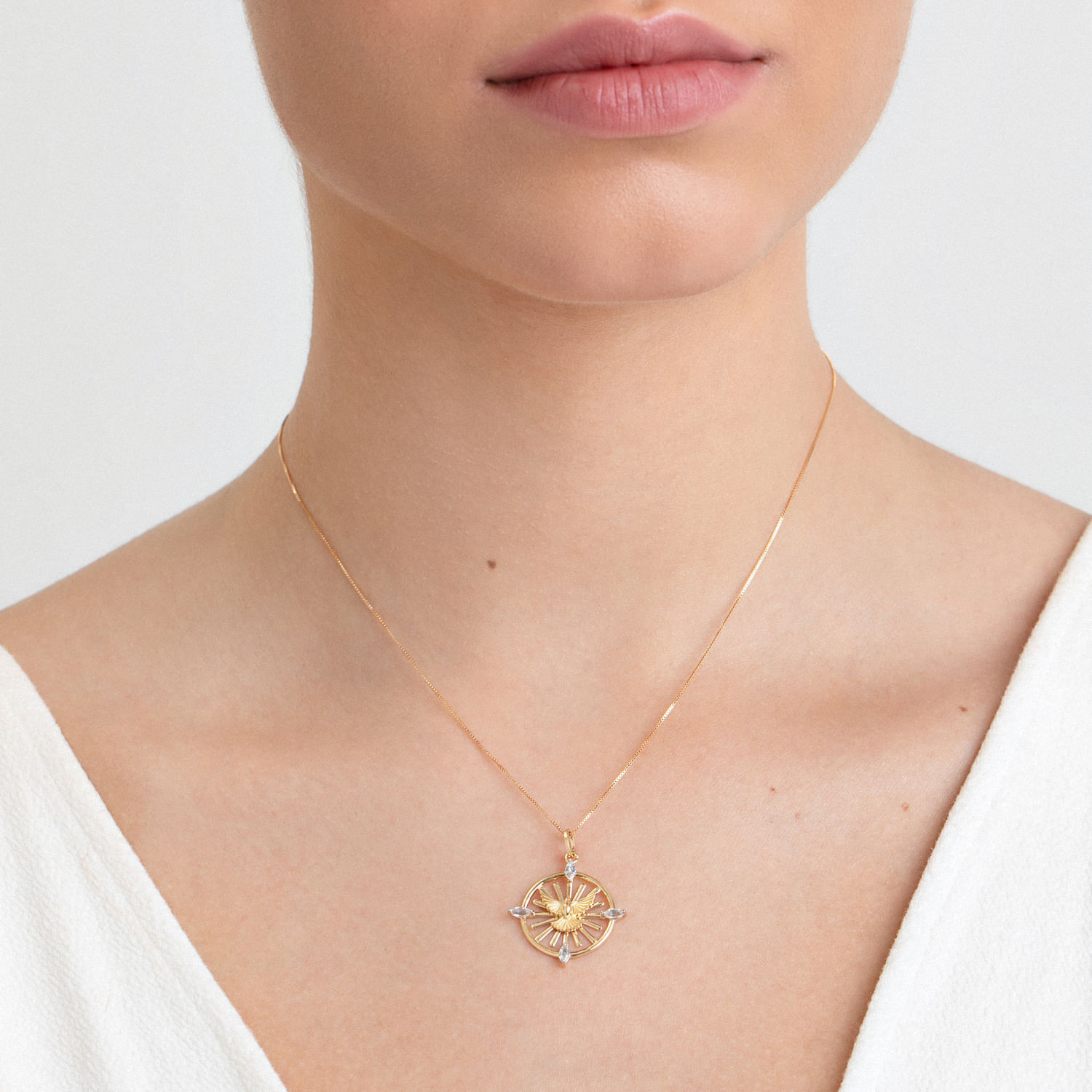 Colar dourado com pingente de sol e detalhes em pedras transparentes, usado por uma pessoa com pele clara e vestindo blusa branca.