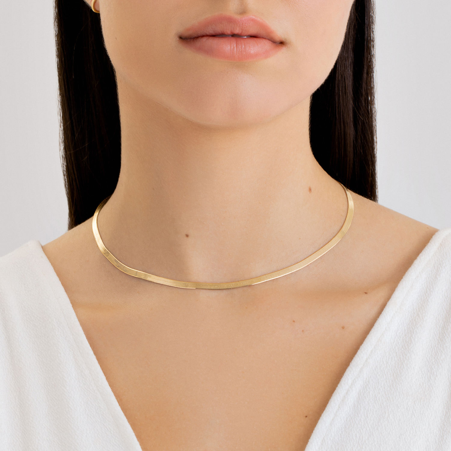 Colar dourado simples e fino sendo usado por uma mulher com pele clara, cabelo castanho escuro e roupa branca.